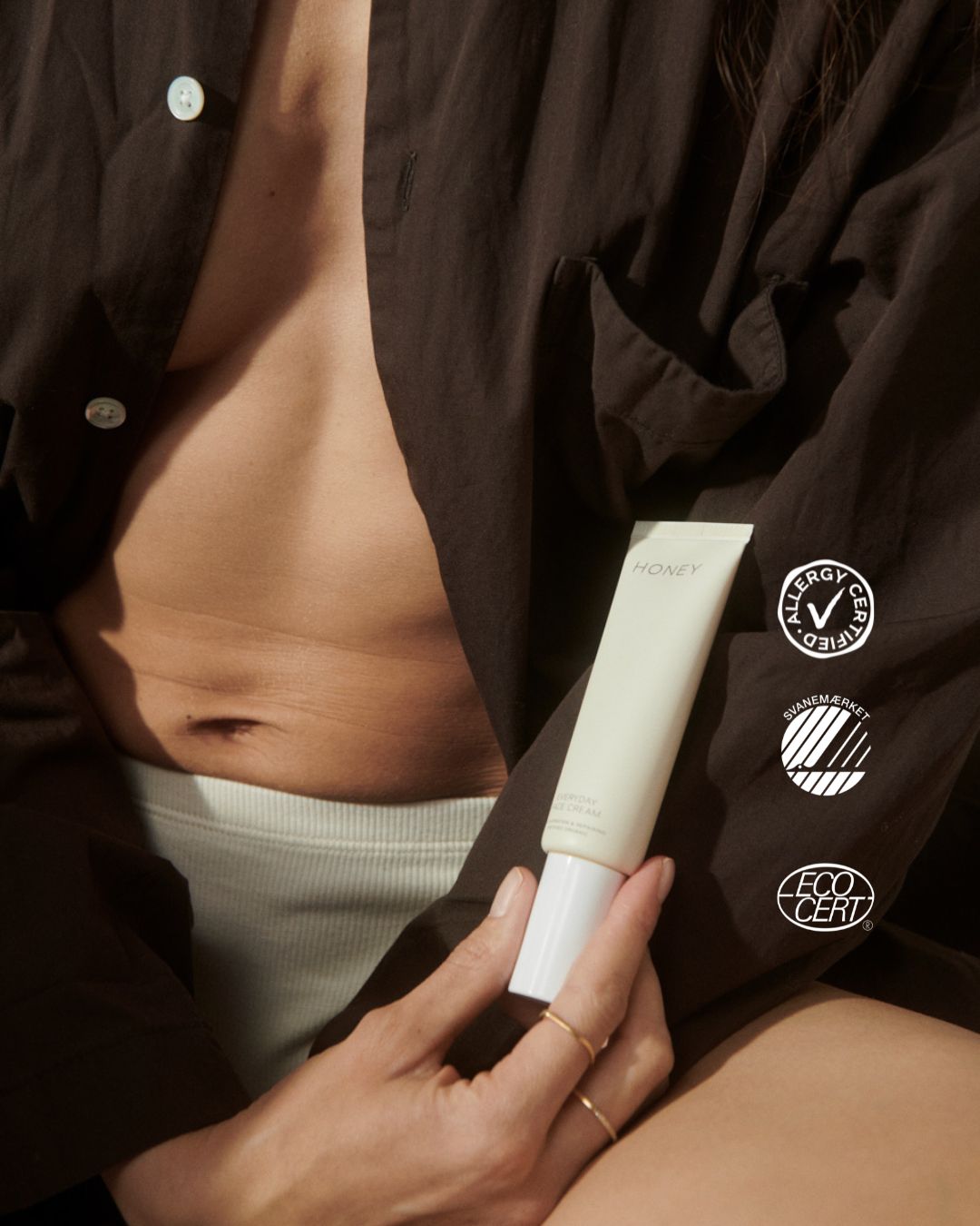 En hånd holder en tube "HONEY everyday face cream" foran en kvinde i en åben brun skjorte. Certificeringsmærker for "ALLERGY CERTIFIED", "SVANEMÆRKET", og "ECO CERT" vises også.