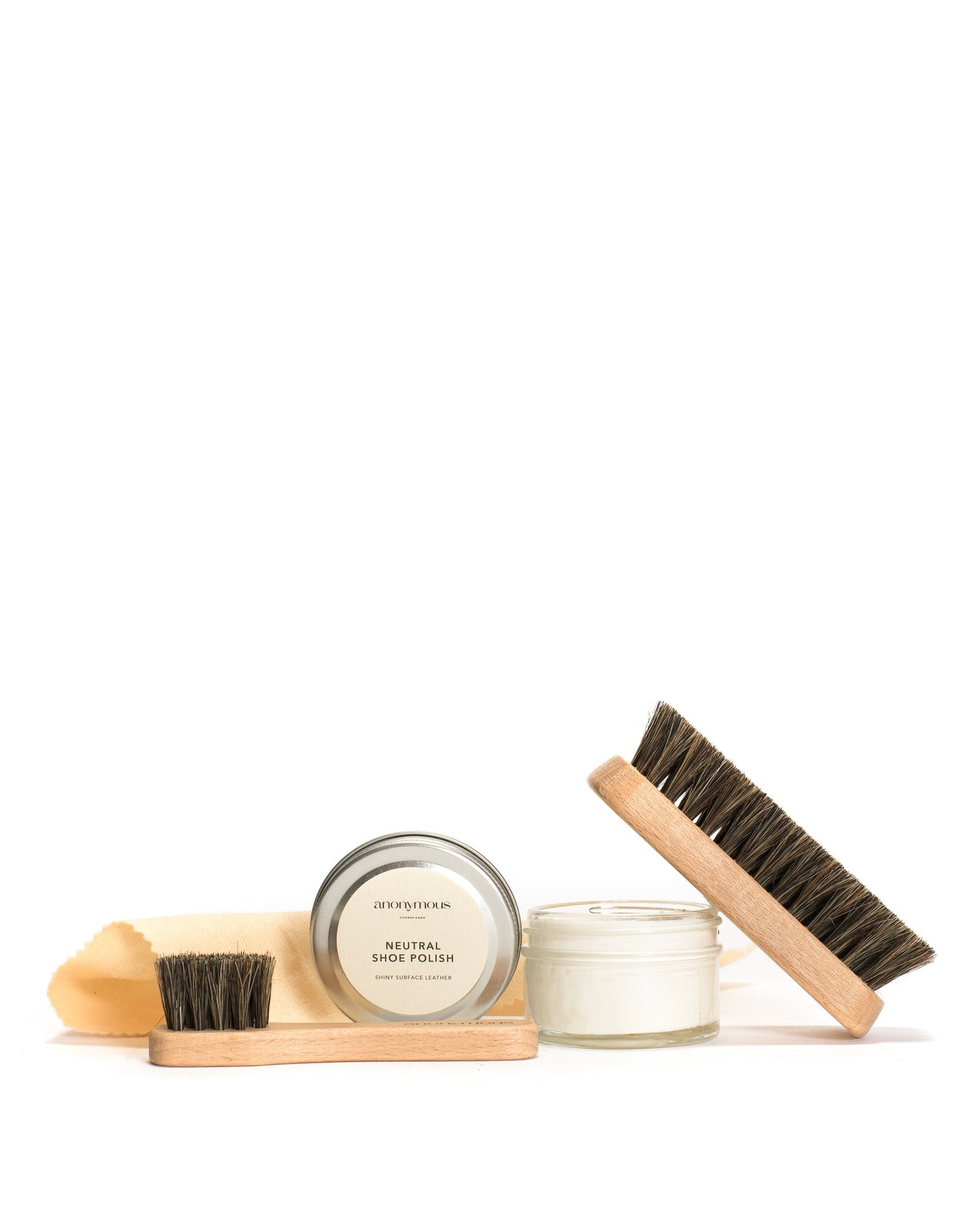 Skoplejeprodukter står på et hvidt underlag. En dåse "anonymous COPENHAGEN NEUTRAL SHOE POLISH SHINY SURFACE LEATHER", en lille glasbeholder, en klud og børster udgør sættet.