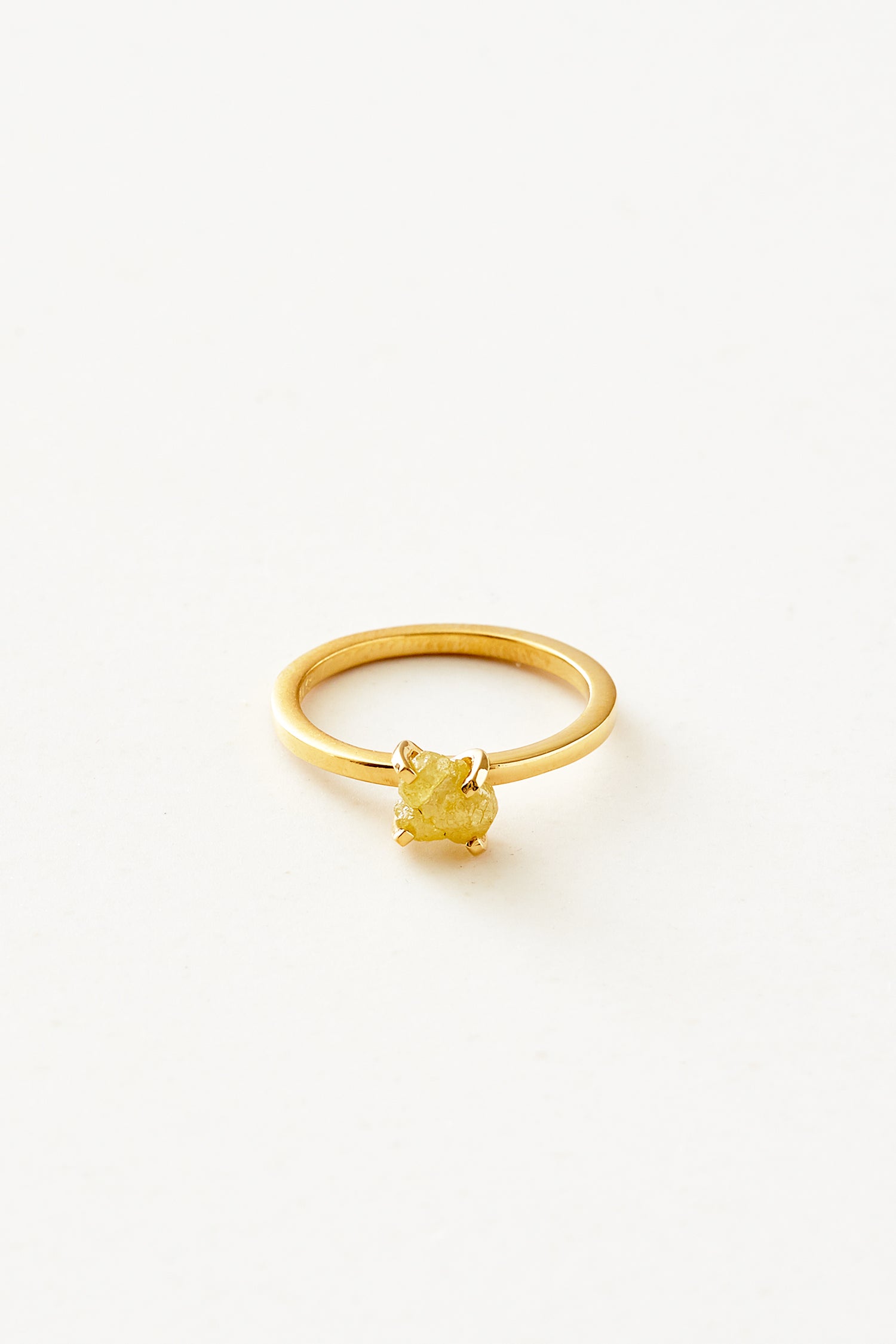 En guldring prydes af en rå, gul diamant, holdt på plads af små kløer. Ringen ligger isoleret på en lys, hvid baggrund.