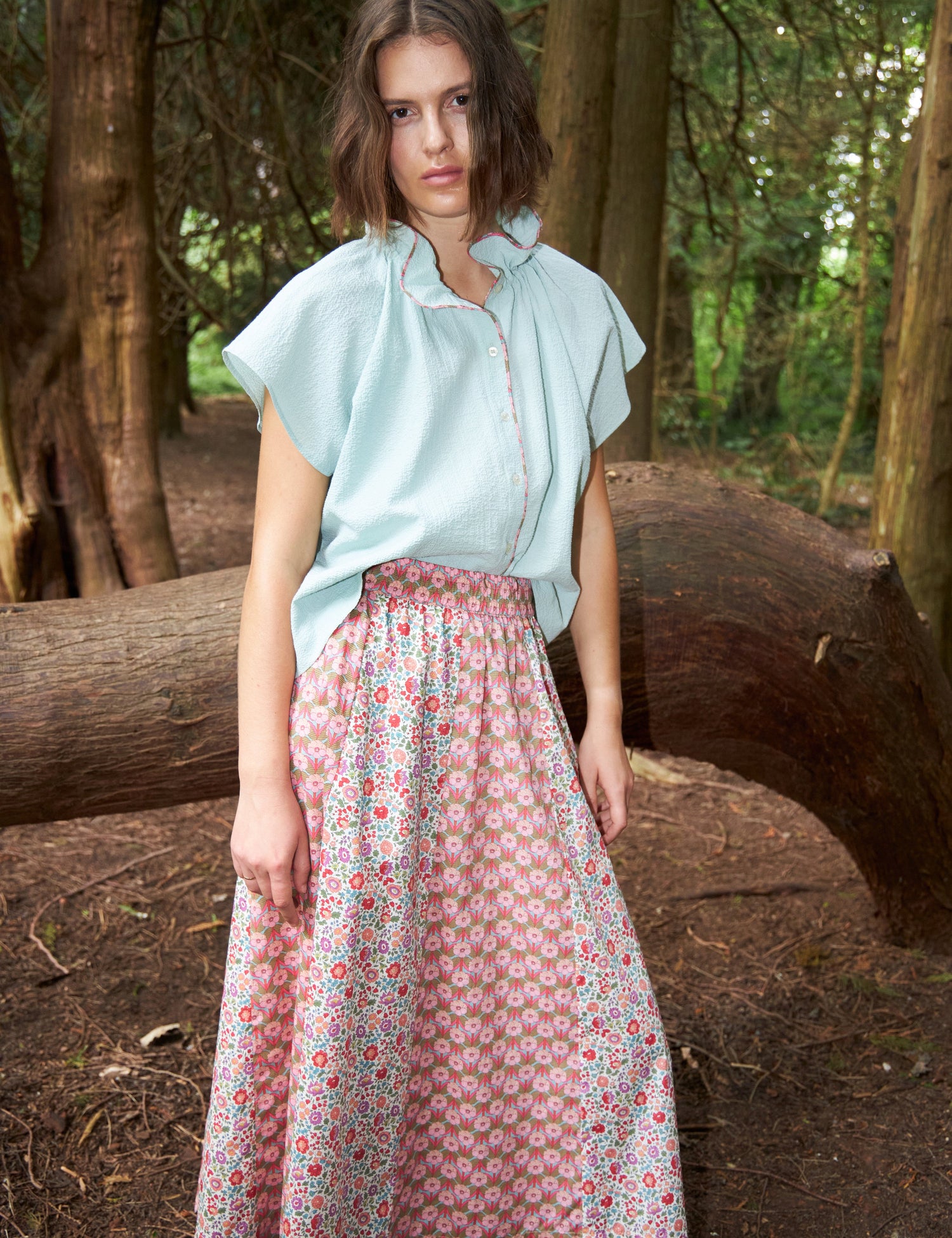 En kvinde står i en skov. Hun er iført en lyseblå bluse med en blomstret nederdel, og kigger direkte på beskueren.
