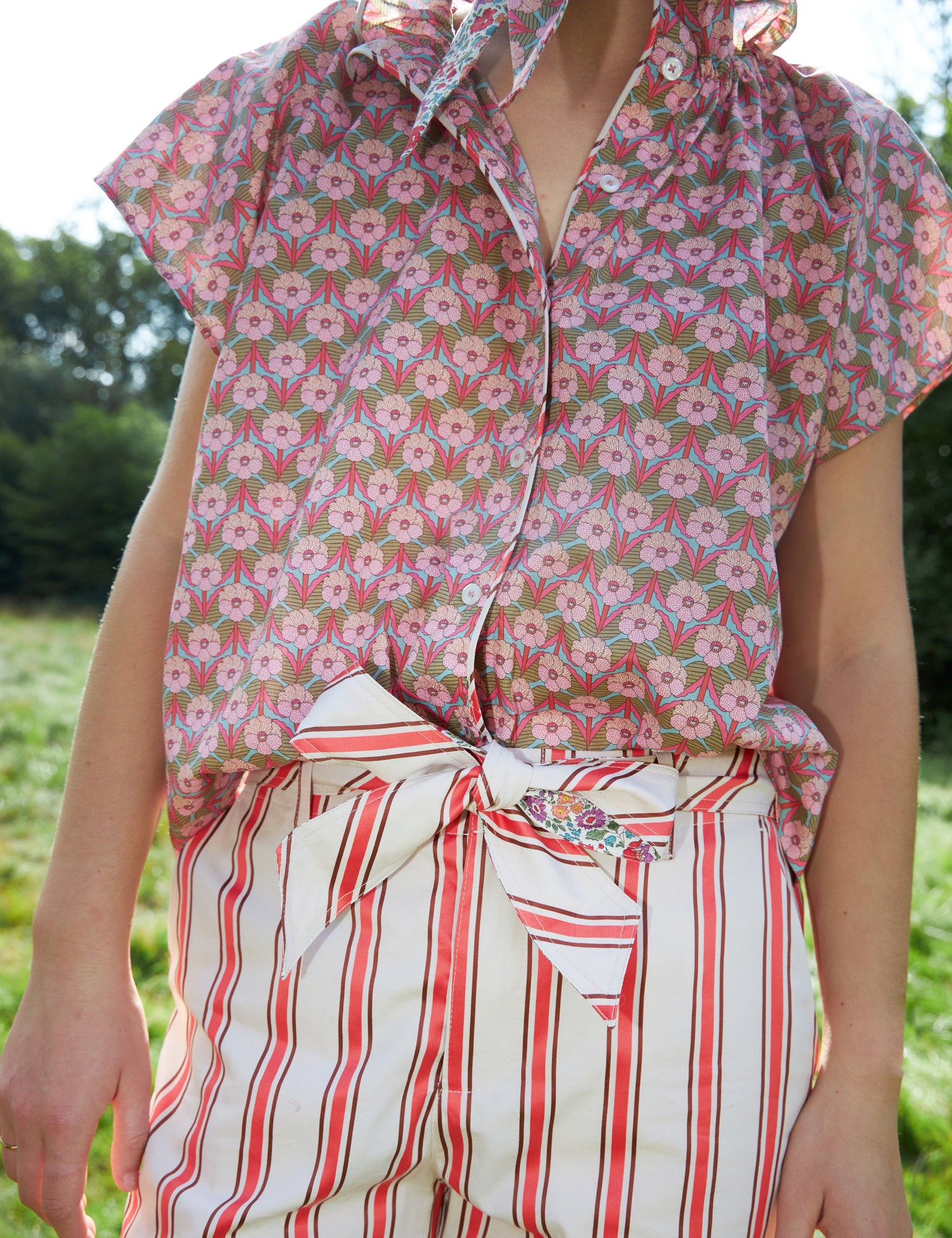 En person bærer en blomstret skjorte og stribede shorts med et bundet bånd i en udendørs setting med grønt græs i baggrunden.