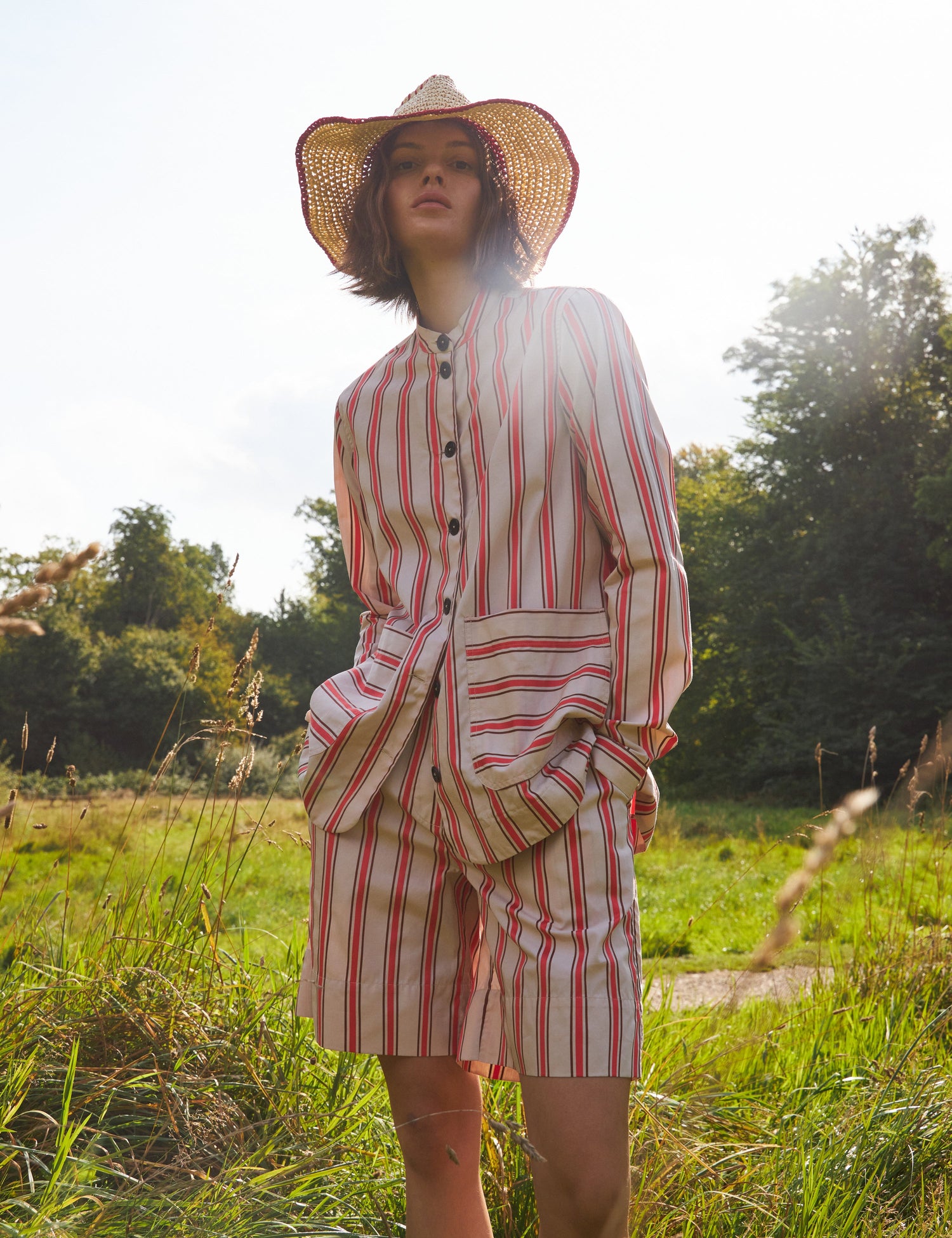 En model poserer i en stribet dragt og stråhat i et grønt græslandskab med træer i baggrunden og solen skinnende ned.