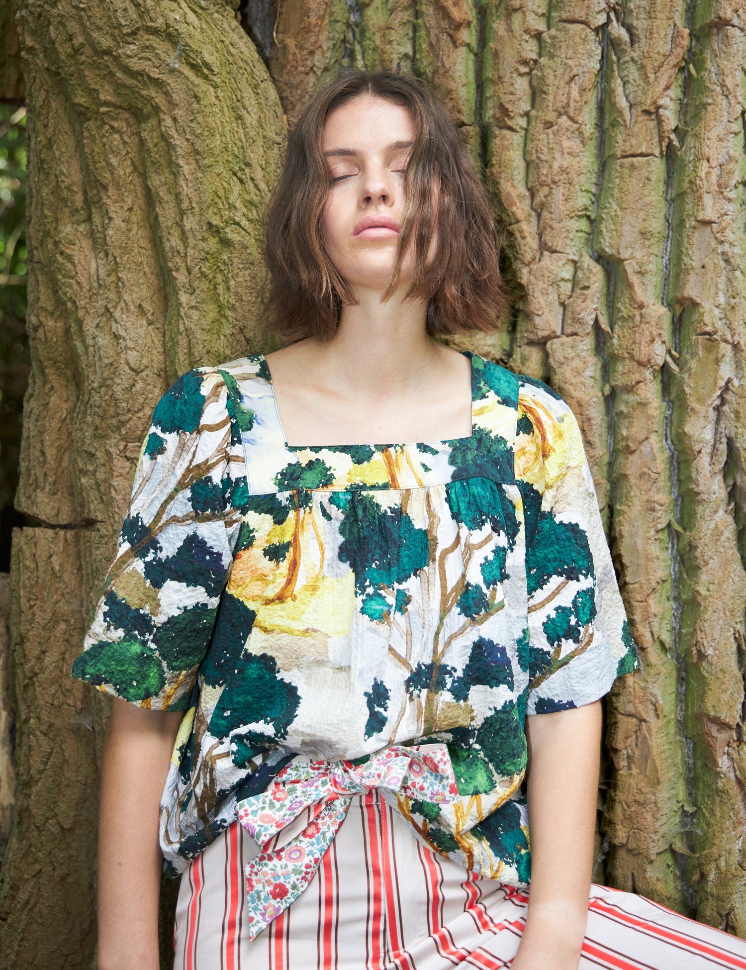 En kvinde med lukkede øjne læner sig mod et træ med bark. Hun bærer en bluse med et malerisk trækronesmønster og stribede bukser med en blomstret sløjfe.