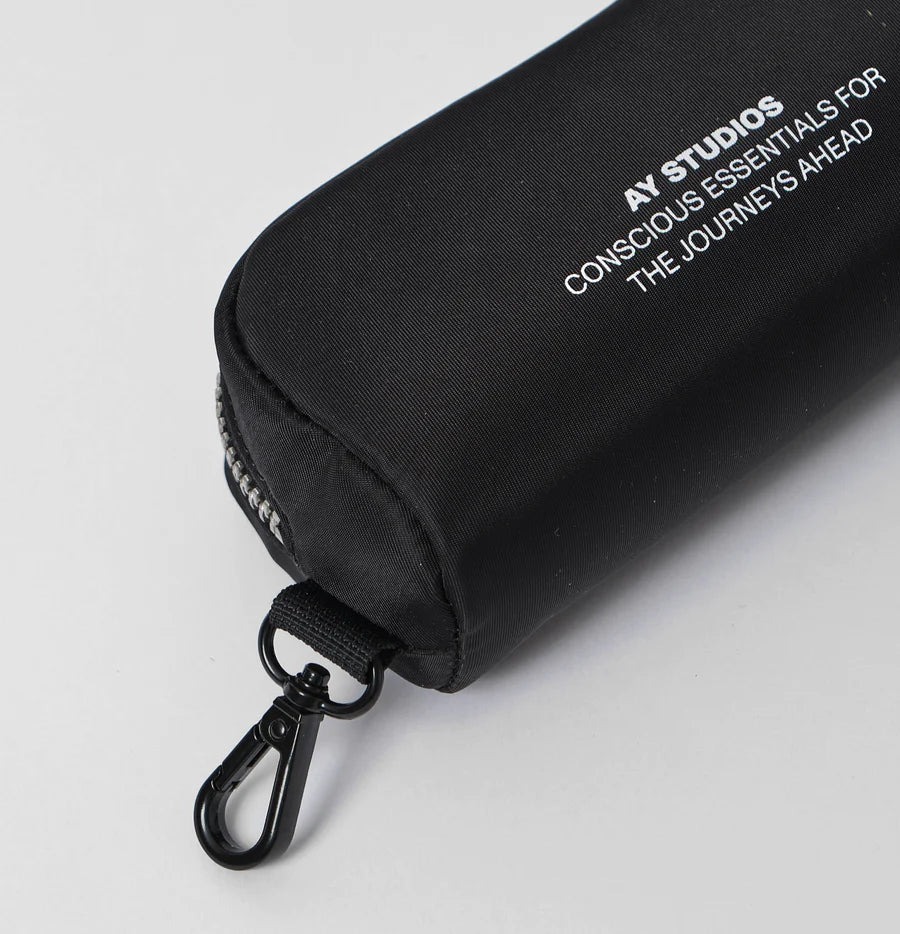 En aflang, sort nylon-etui ligger på en hvid overflade. En sølvfarvet lynlås ses, og etuiet er mærket "AY STUDIOS CONSCIOUS ESSENTIALS FOR THE JOURNEYS AHEAD". En sort karabinhage er fastgjort.