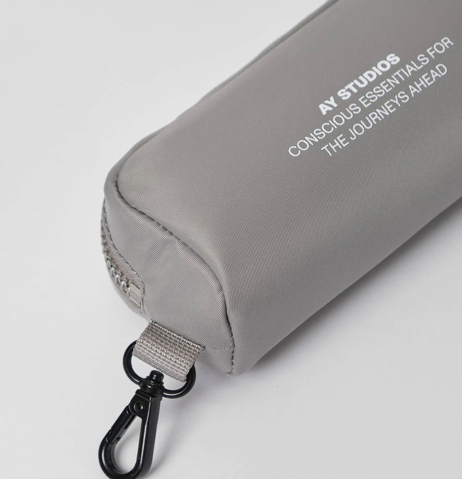 En grå taske med lynlås og en sort karabinhage. Teksten "AY STUDIOS CONSCIOUS ESSENTIALS FOR THE JOURNEYS AHEAD" er trykt på siden.