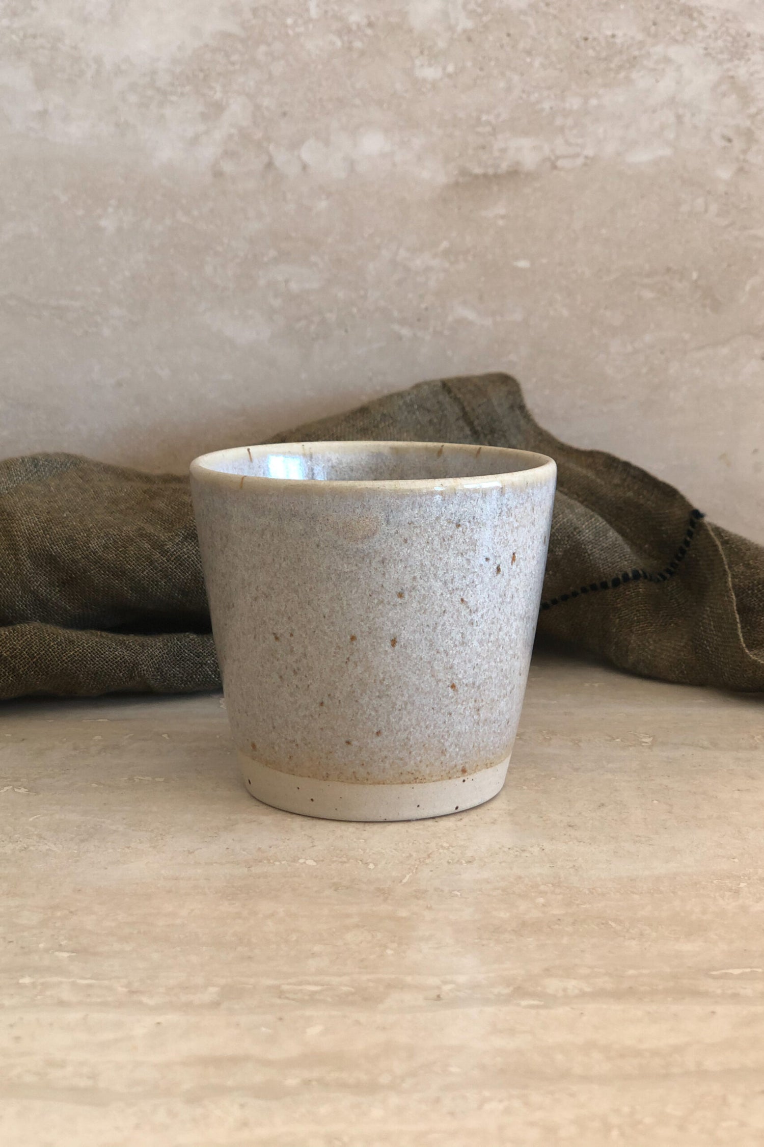 En keramisk kop står stille på en beige marmoroverflade. En foldet, gråbrun tekstil baggrund danner en kontrast til koppen.