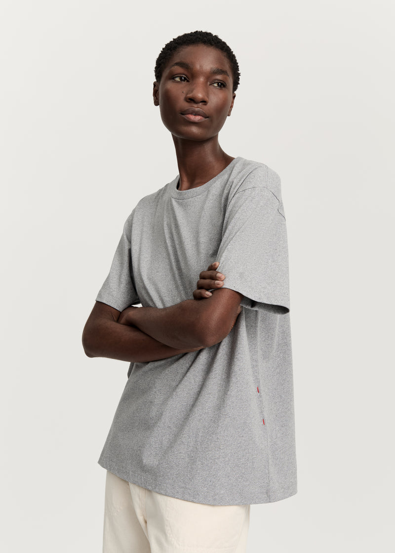 En model poserer i en grå T-shirt med krydsede arme foran en hvid baggrund. T-shirten er kombineret med lyse bukser.