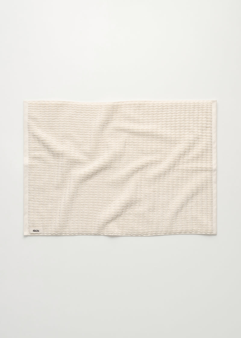 Et cremefarvet vaffelmønstret håndklæde ligger fladt på en hvid baggrund. Et lille sort mærke med hvid skrift "aiaiai" er synligt i nederste venstre hjørne.