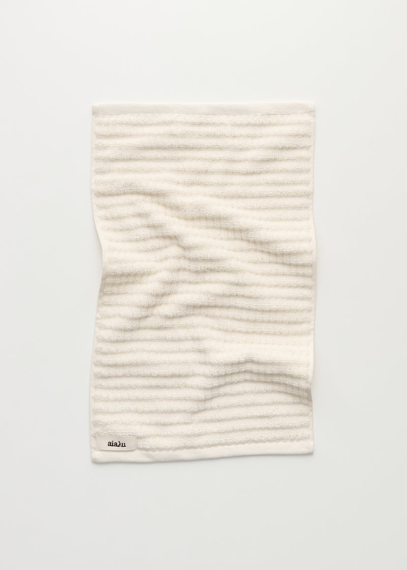 Et stribet, cremefarvet håndklæde ligger foldet på en hvid overflade. Etiket nederst til venstre viser teksten "aiaLu".