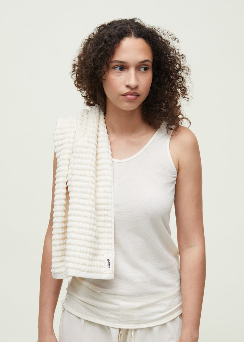 En kvinde med krøllet hår har et stribet håndklæde over skulderen. Hun er iført en hvid tanktop i et lyst studie. Håndklædet har en etiket med teksten "aia:au".