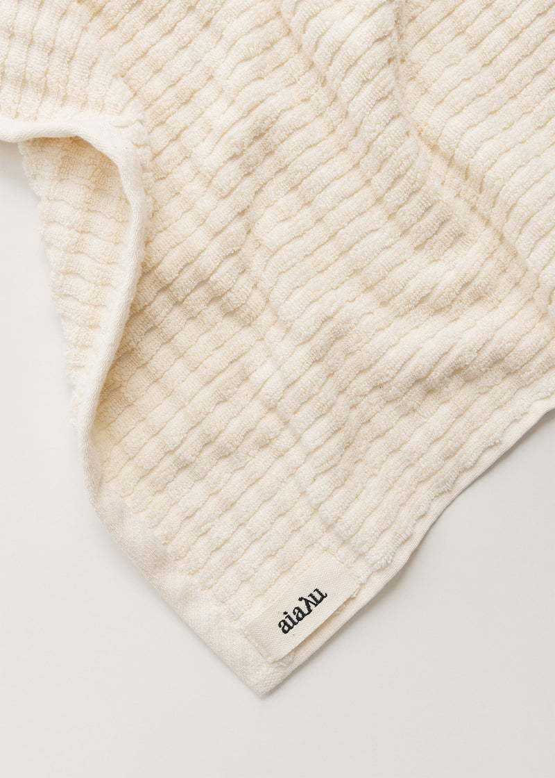 En cremefarvet vaskeklud med et bølget mønster ligger foldet på en hvid overflade. Etiketten i nederste hjørne viser teksten "aiaLu".