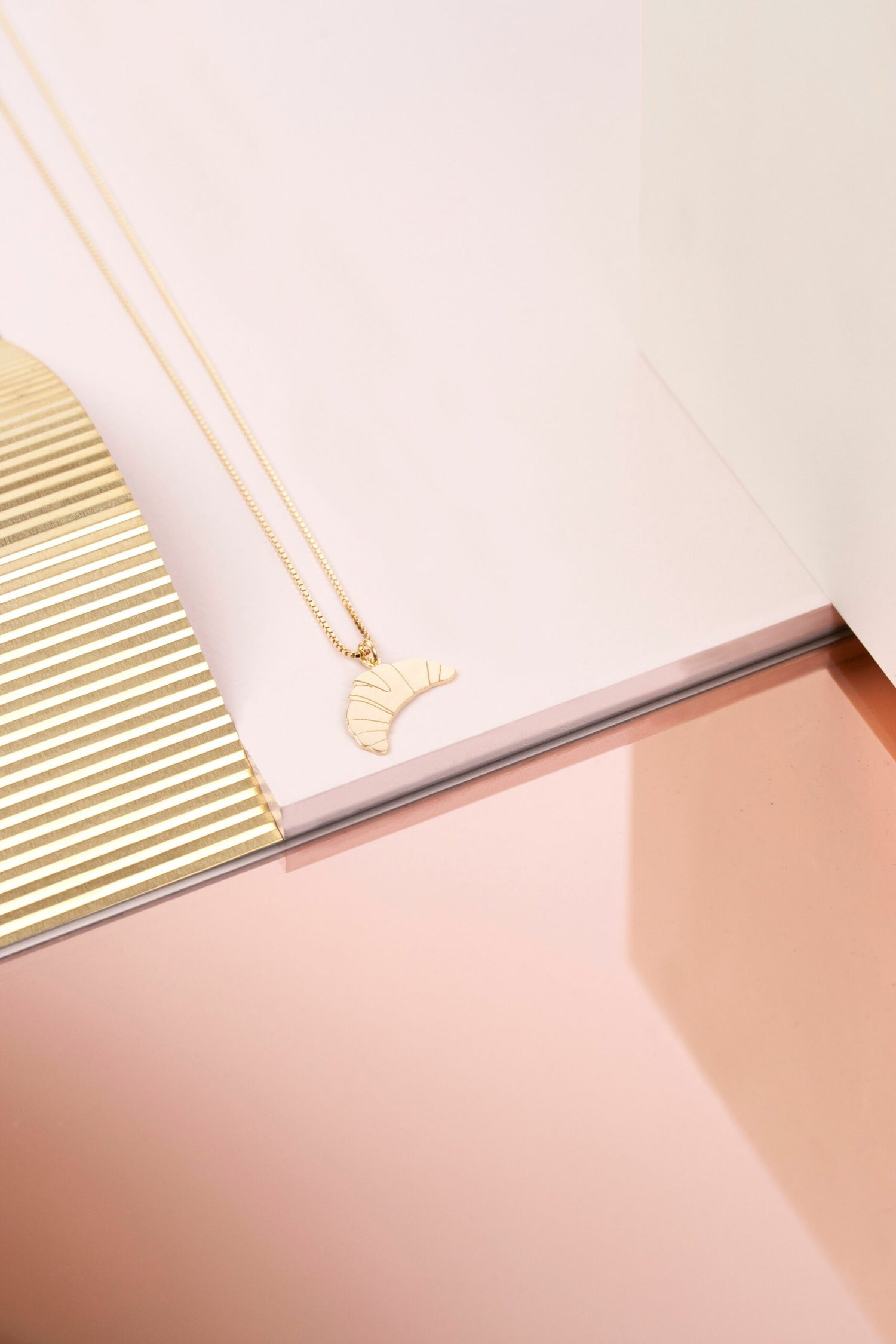 Et gyldent croissant-vedhæng ligger på en åben bog. Vedhænget er fastgjort til en fin, gylden kæde. Bogen og en dekoration har en sart, lyserød farve.