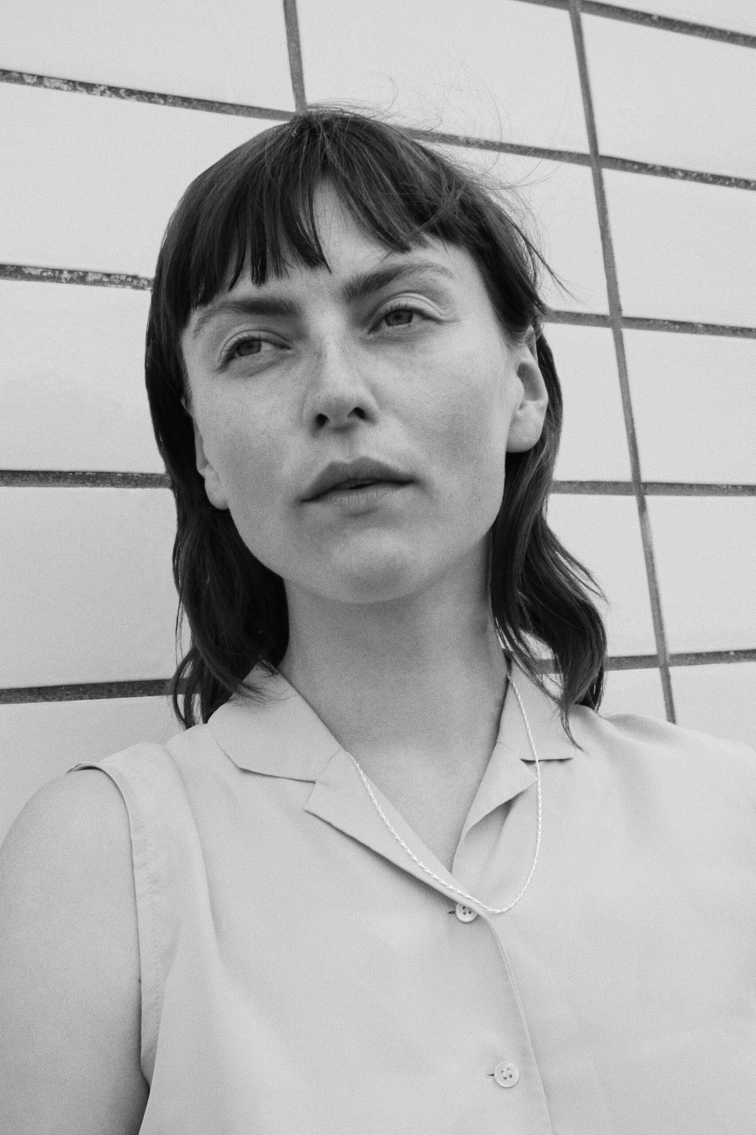 En kvinde med pandehår ser opad, iført en skjorte med knapper og en fin halskæde. Hun står foran en hvid flisevæg.