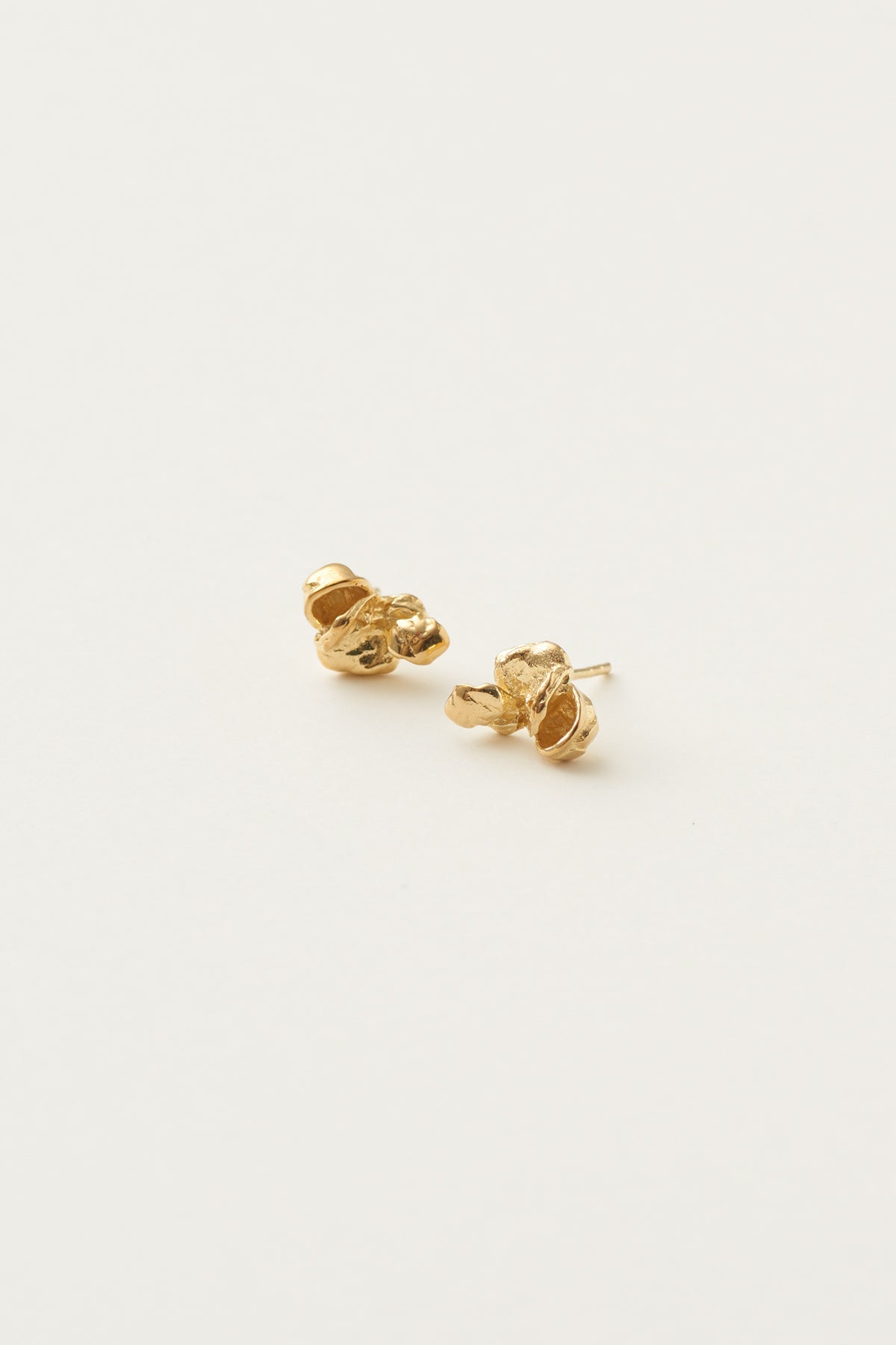 To gyldne øreringe ligger på en hvid overflade. Øreringene har en organisk, klumpet form og en tekstureret overflade.