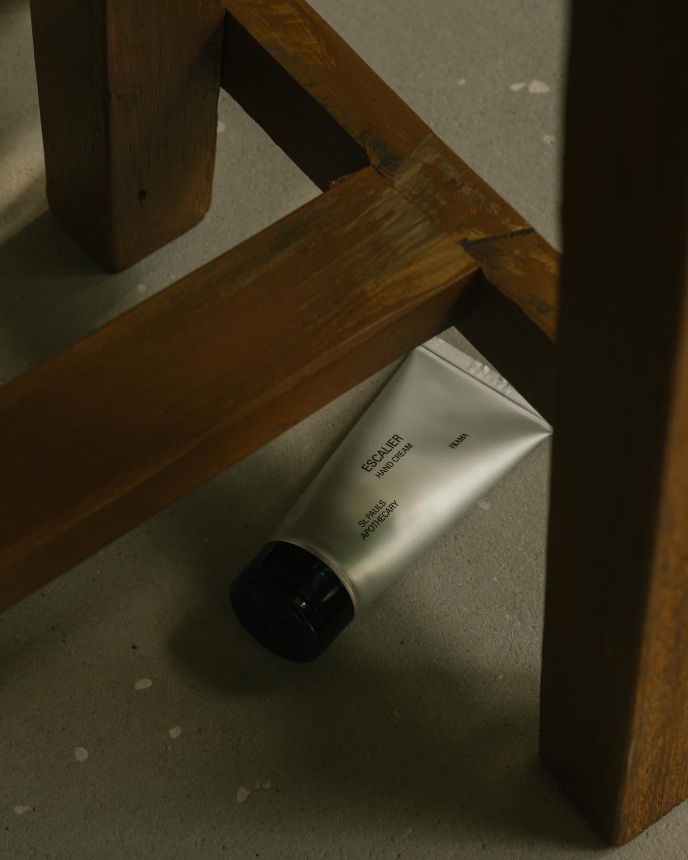 En tube håndcreme ligger på et betongulv, delvist under en træstolsben. Tuben er mærket med "ESCALIER HAND CREAM, FRAMA, St. PAULS APOTHECARY".