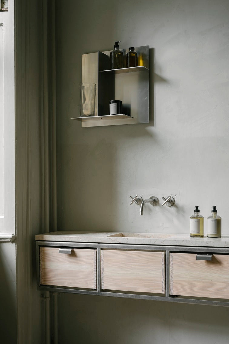 Et badeværelsesmiljø viser en vask med et kabinet med træskuffer og en kromhylde med glas og flasker mod en simpel, tekstureret væg.