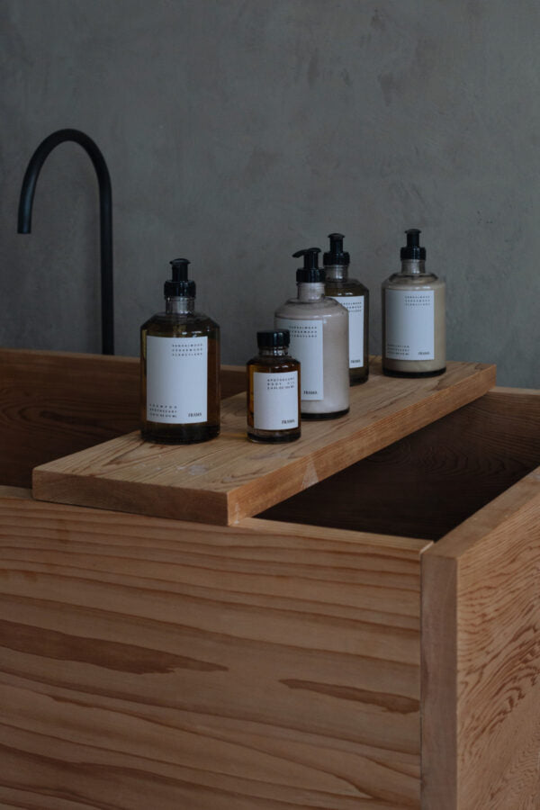 Sæber og lotion står på en træplade over et trækar. Teksten på etiketterne lyder: "SANDALWOOD CARDAMOM SERUM/LOTION, FRAMA."