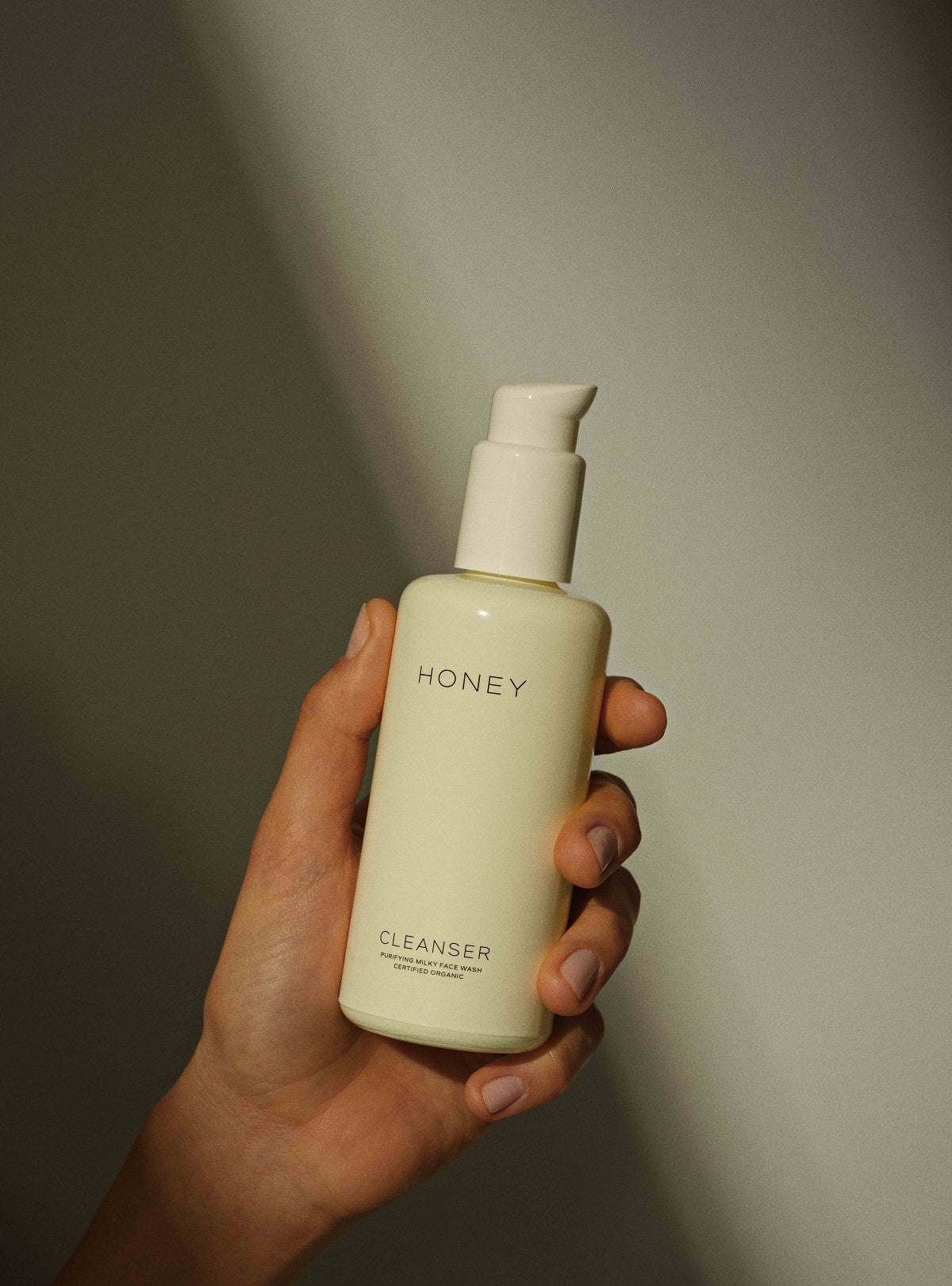 En hånd holder en creme-farvet flaske med pumpetop. Teksten "HONEY CLEANSER PURIFYING MILKY FACE WASH CERTIFIED ORGANIC" står på flasken. Bagrunden er en simpel lys væg.