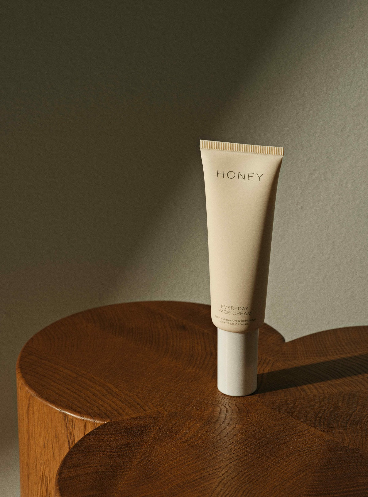 En tube "HONEY EVERYDAY FACE CREAM. DEEP HYDRATION & REPAIRING. CERTIFIED ORGANIC" står oprejst på et træbord. Tuben er beige med hvid låg. Baggrunden er en ensfarvet væg.