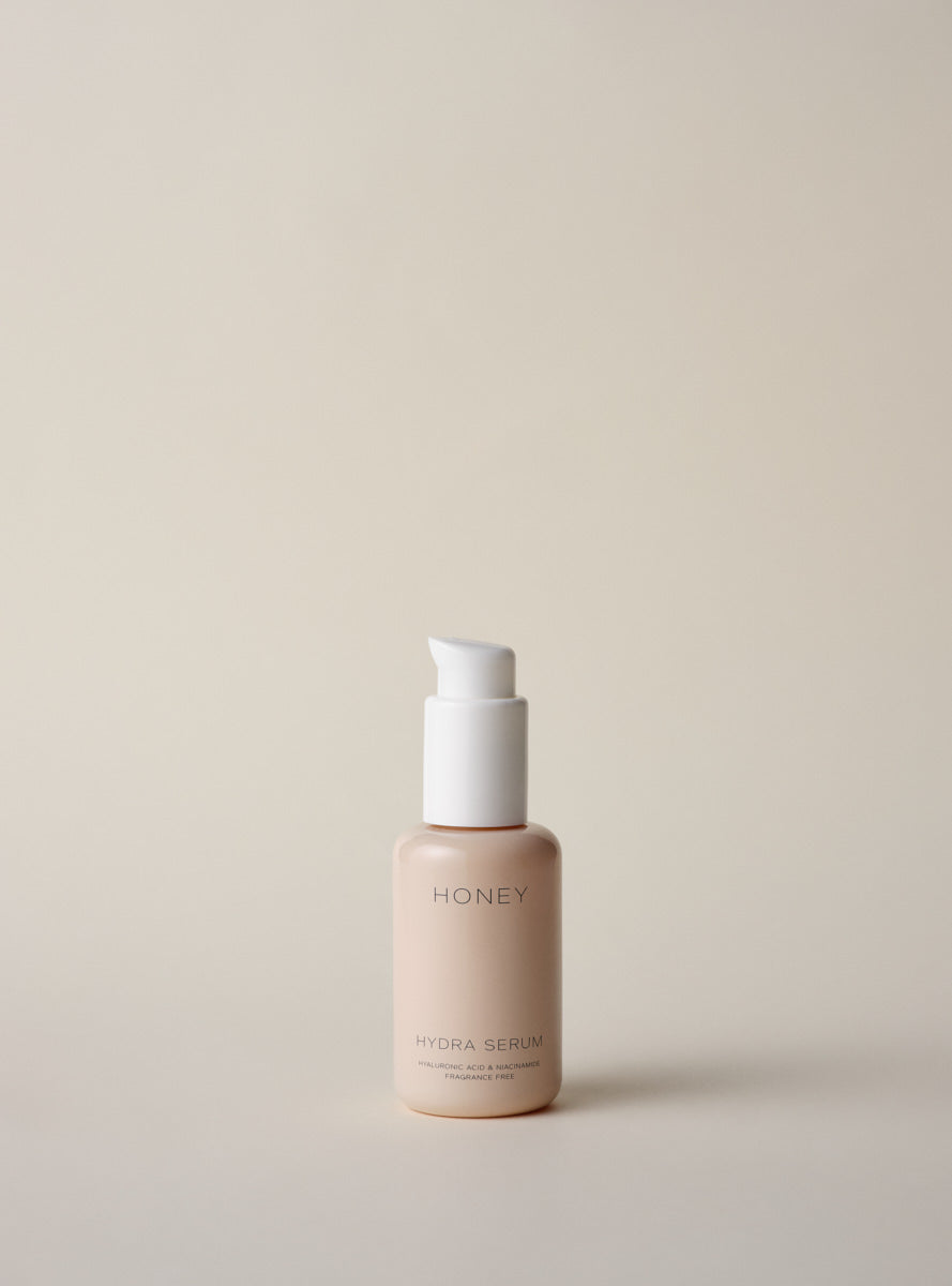En flaske med en hvid pumpe står på en lys beige baggrund. Flasken er mærket med "HONEY", "HYDRA SERUM", "HYALURONIC ACID & NIACINAMIDE" og "FRAGRANCE FREE".