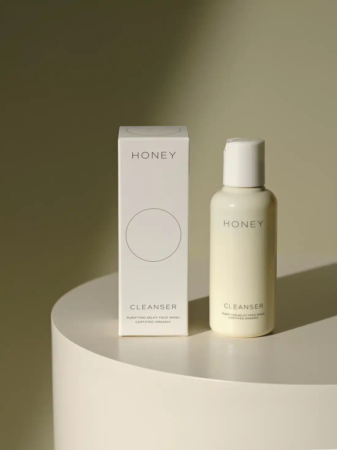 En hvid flaske og en kasse, begge mærket "HONEY CLEANSER PURIFYING MILKY FACE WASH CERTIFIED ORGANIC", står på en hvid piedestal mod en olivengrøn baggrund.