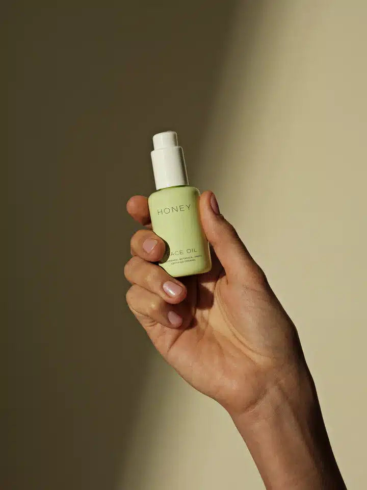 En hånd holder en flaske "HONEY FACE OIL ORGANIC, BOTANICAL, FRESH, CRAFTED, ORGANIC" mod en neutral baggrund. Flasken er lysegrøn med en hvid pumpe.