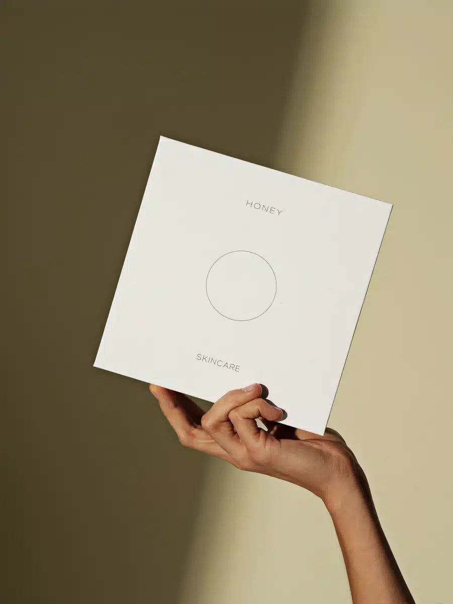 En hvid æske med teksten "HONEY" og "SKINCARE" holdes af en hånd mod en ensfarvet beige baggrund med skygger. Æsken har et simpelt, minimalistisk design med en cirkel i midten.