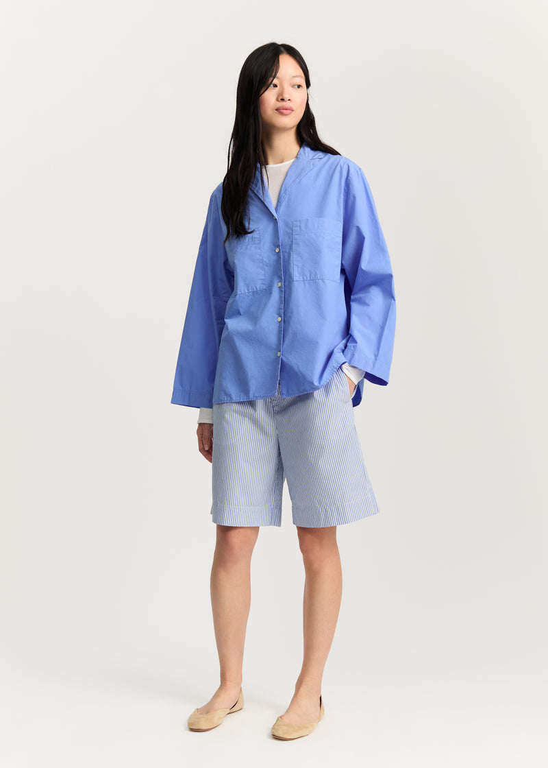 En kvinde står i et hvidt studie, iført en blå skjorte med lange ærmer, stribede shorts og flade sko, og ser ud til højre.