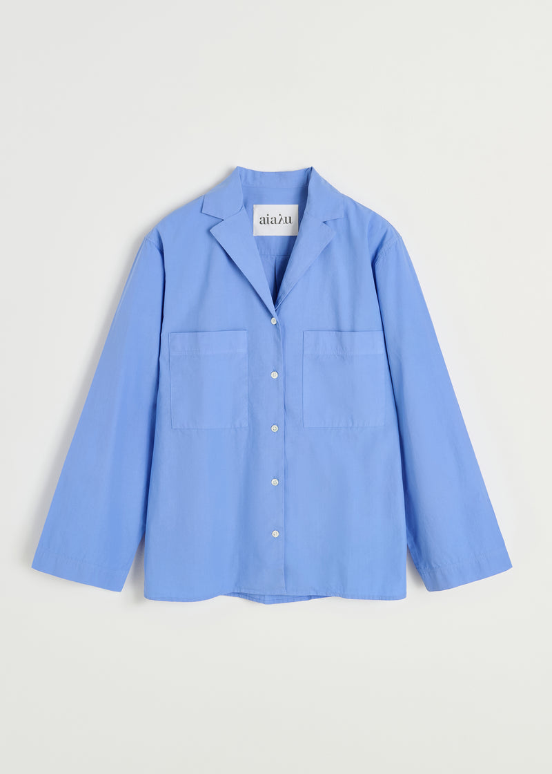 En lyseblå skjorte med lange ærmer, brystlommer og en "aiaïu"-etiket i kraven ligger fladt på en hvid baggrund.