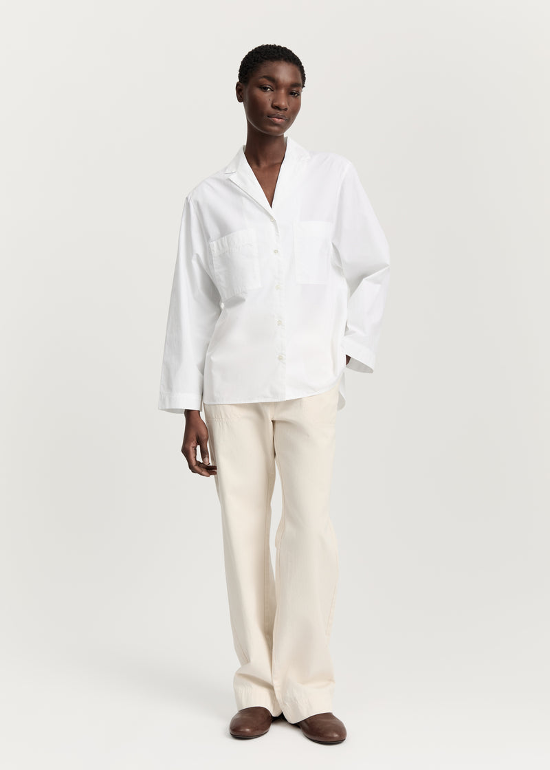 En kvinde poserer i en hvid skjorte med lange ærmer og lyse bukser foran en hvid baggrund. Hun har brune sko på.