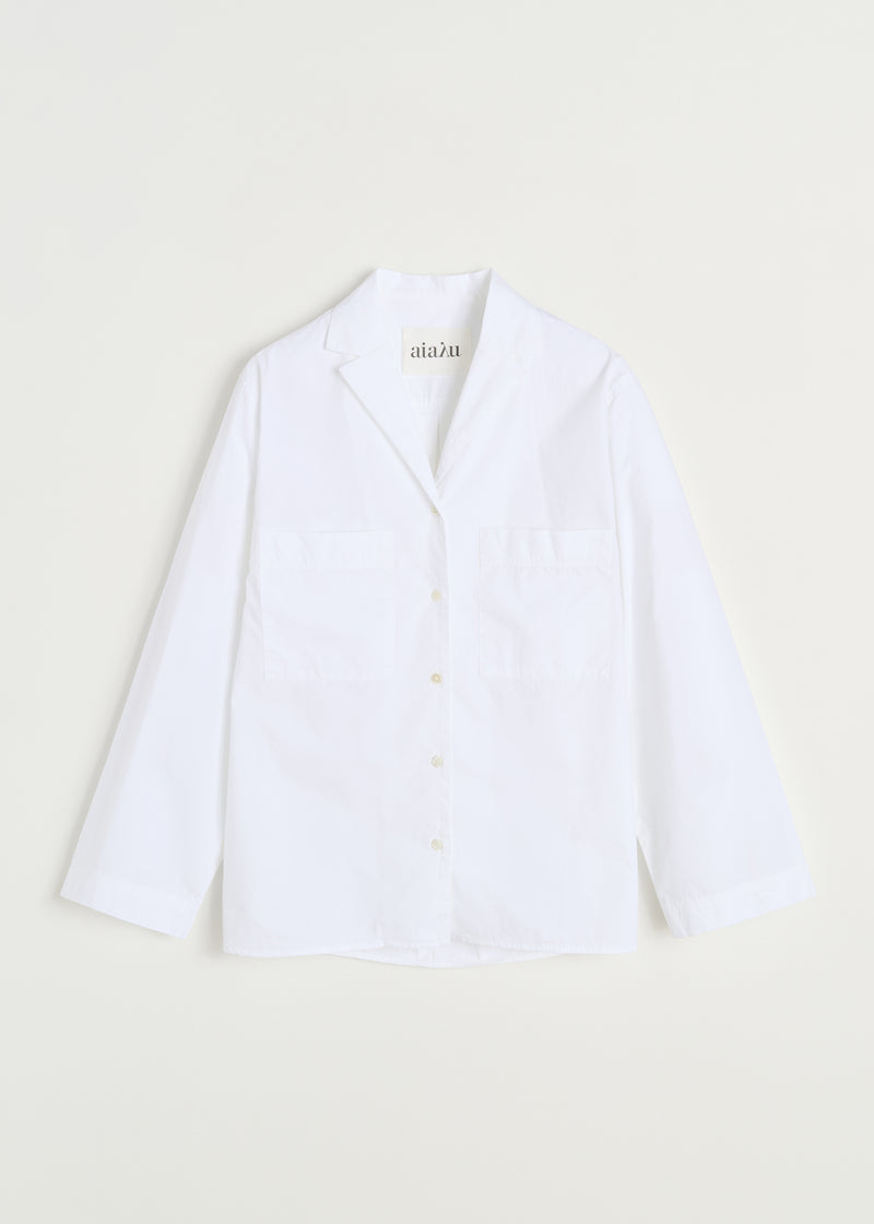 En hvid skjorte med lange ærmer, to lommer og en åben krave ligger fladt mod en off-white baggrund. Der står "aiaìu" på etiketten.
