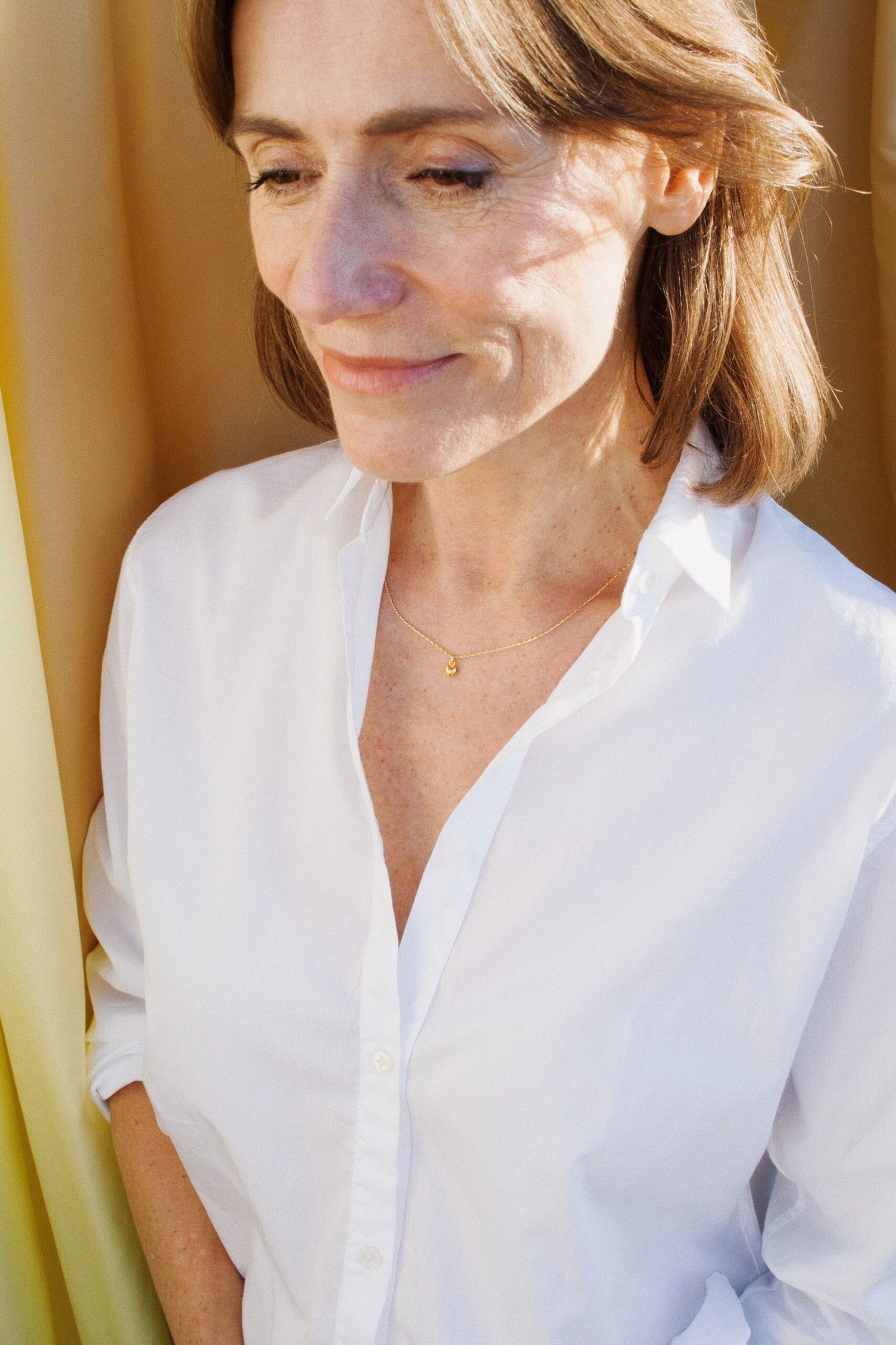 En kvinde med brunt hår og hvid skjorte smiler let. Hun bærer en guldkæde med et lille vedhæng foran en gul baggrund.