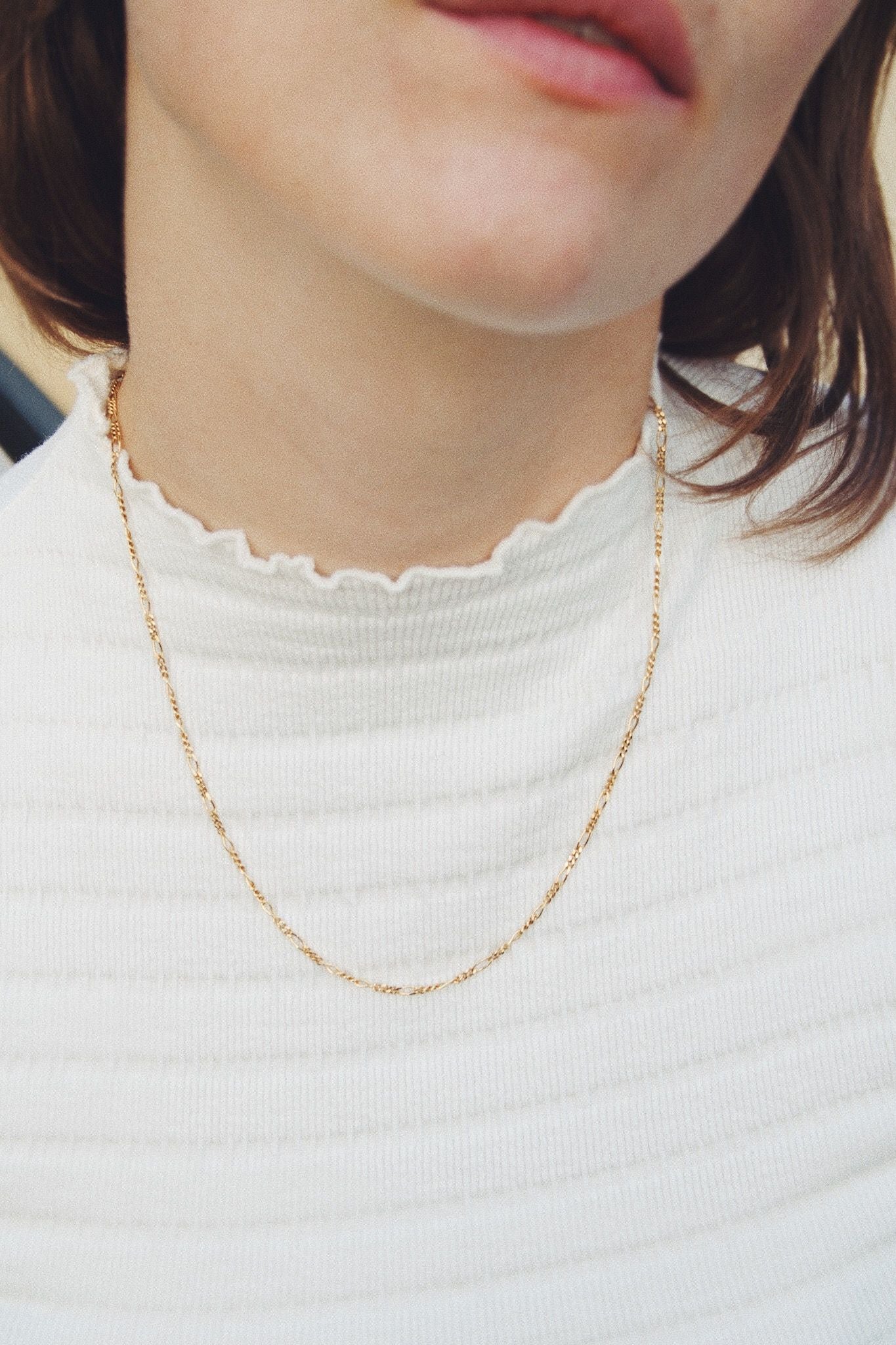 Et guldfarvet halskæde hænger om halsen på en kvinde, der bærer en hvid trøje med flæser ved halsudskæringen. Hendes læber er synlige foroven.