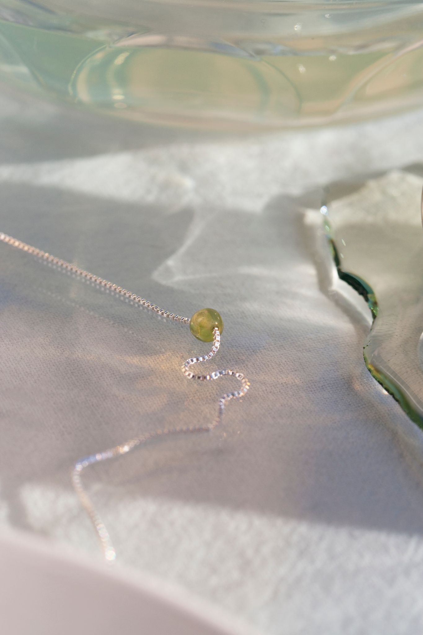 Et sølvsmykke med en grøn perle ligger på en hvid overflade nær et glasobjekt med vand. Lyset skaber bløde skygger på overfladen.