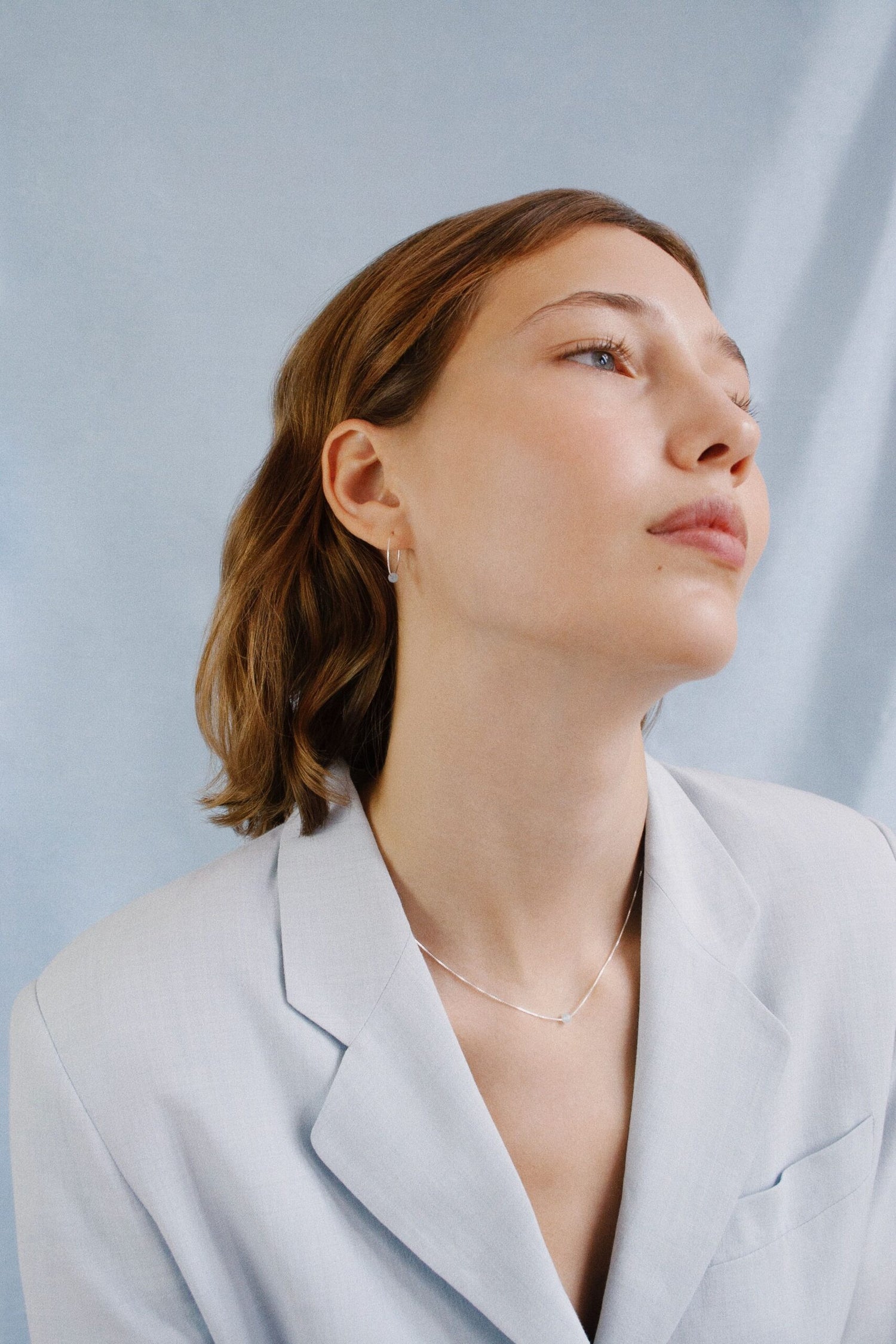 En kvinde med kort brunt hår kigger opad. Hun bærer en lyseblå blazer og en fin sølvkæde mod en lyseblå baggrund.