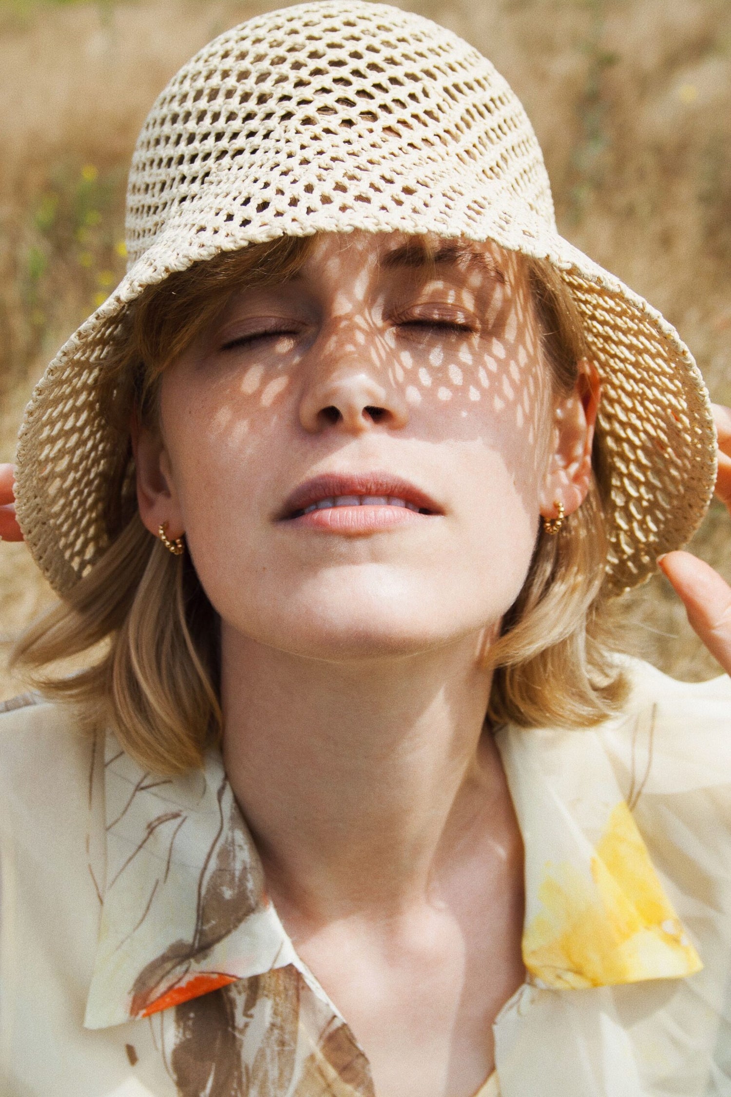 En kvinde med lukkede øjne bærer en hat, der kaster et mønster af lys og skygge over hendes ansigt. Hun er udendørs i et felt af tørt græs.