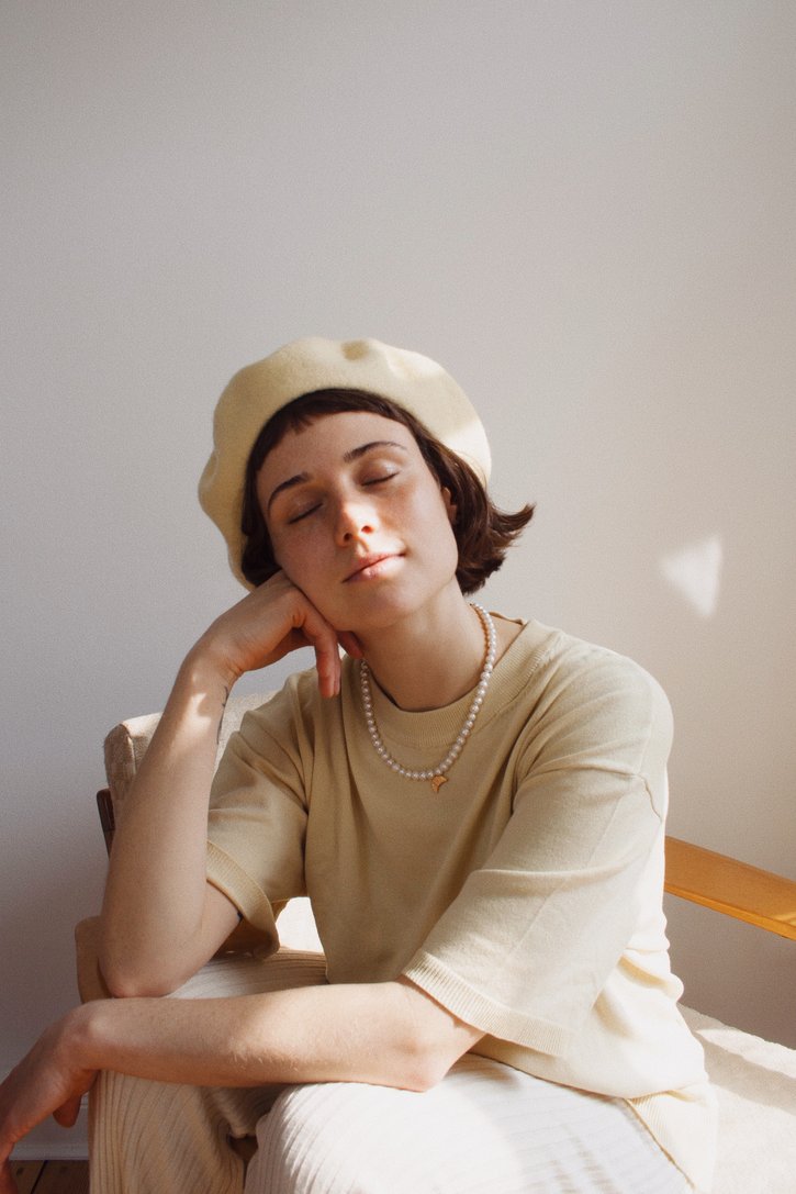 En kvinde med lukket øjne hviler sit hoved i hånden, iført en baret og perlekæde. Hun sidder i en stol mod en neutral væg i naturligt lys.