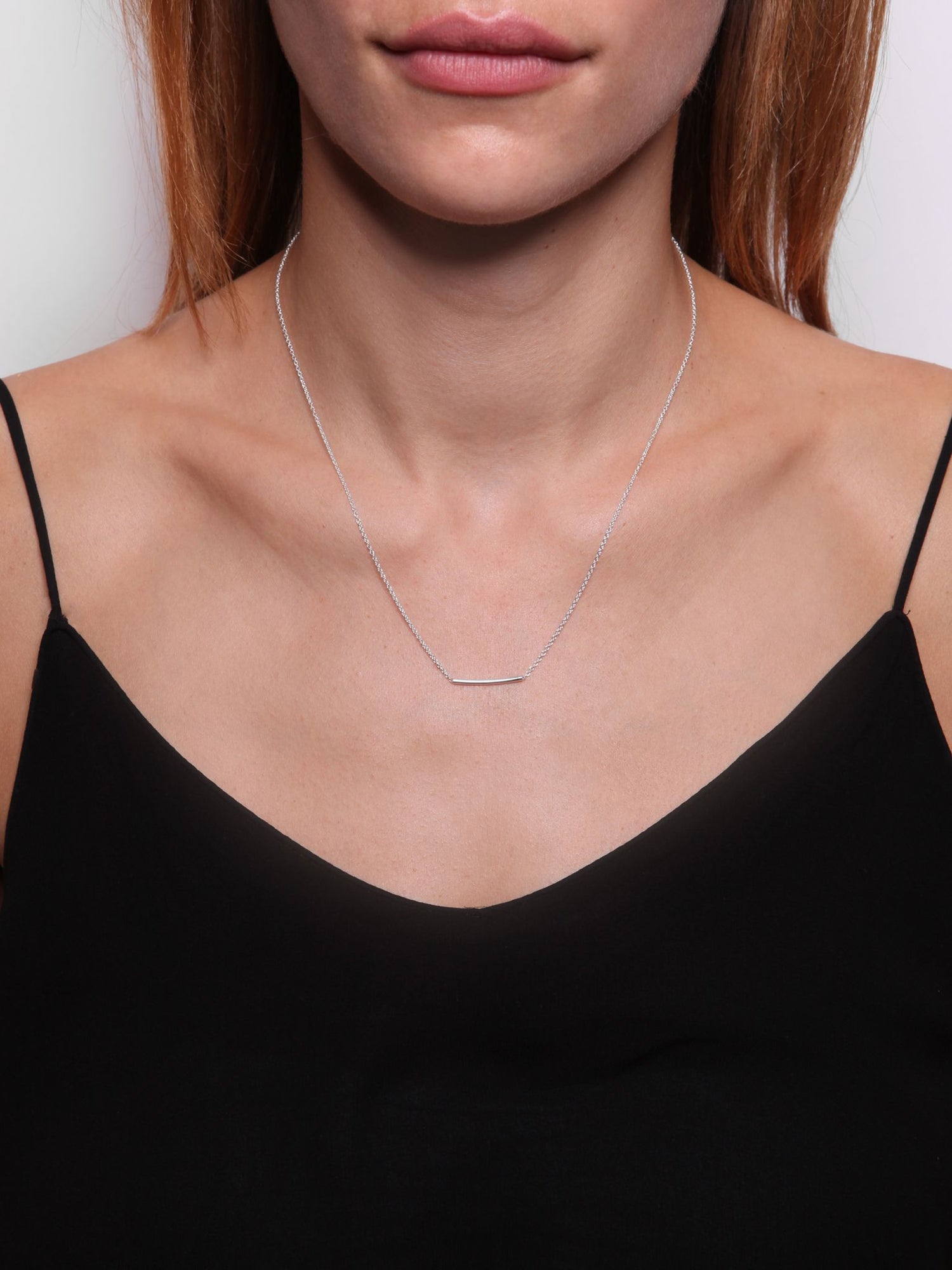 En kvinde bærer en tynd sølvkæde med en diskret, vandret sølvstang ved halsudskæringen af en sort tanktop foran en hvid baggrund.