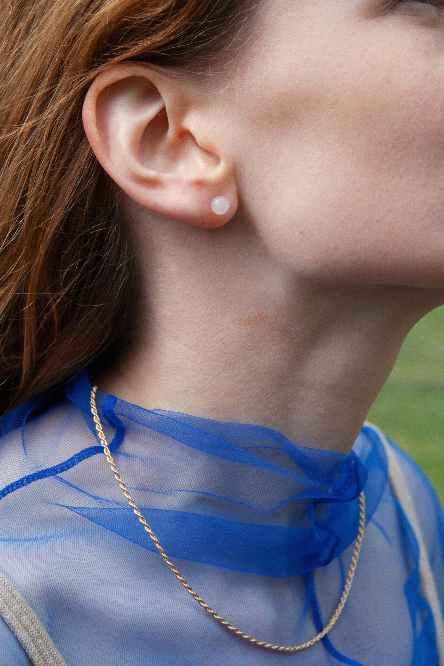 Nærbillede af en kvindes ansigt, hvor en rund, hvid ørering sidder i øret. En guldhalskæde hænger foran en blå tyltop, med grøn baggrund.