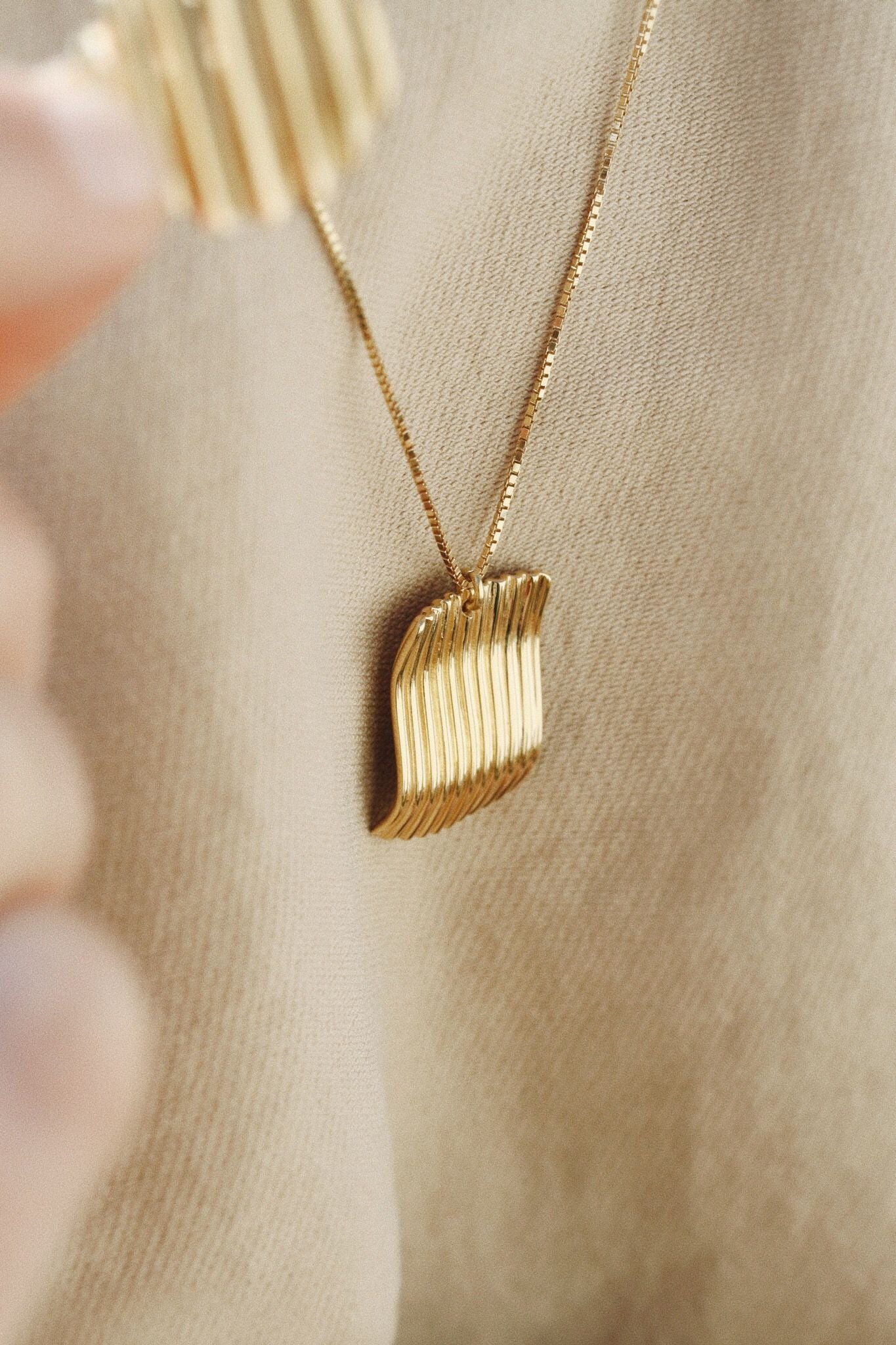Et guldfarvet halskæde med et abstrakt vedhæng hviler mod et beige stof. Finger ses i venstre side.