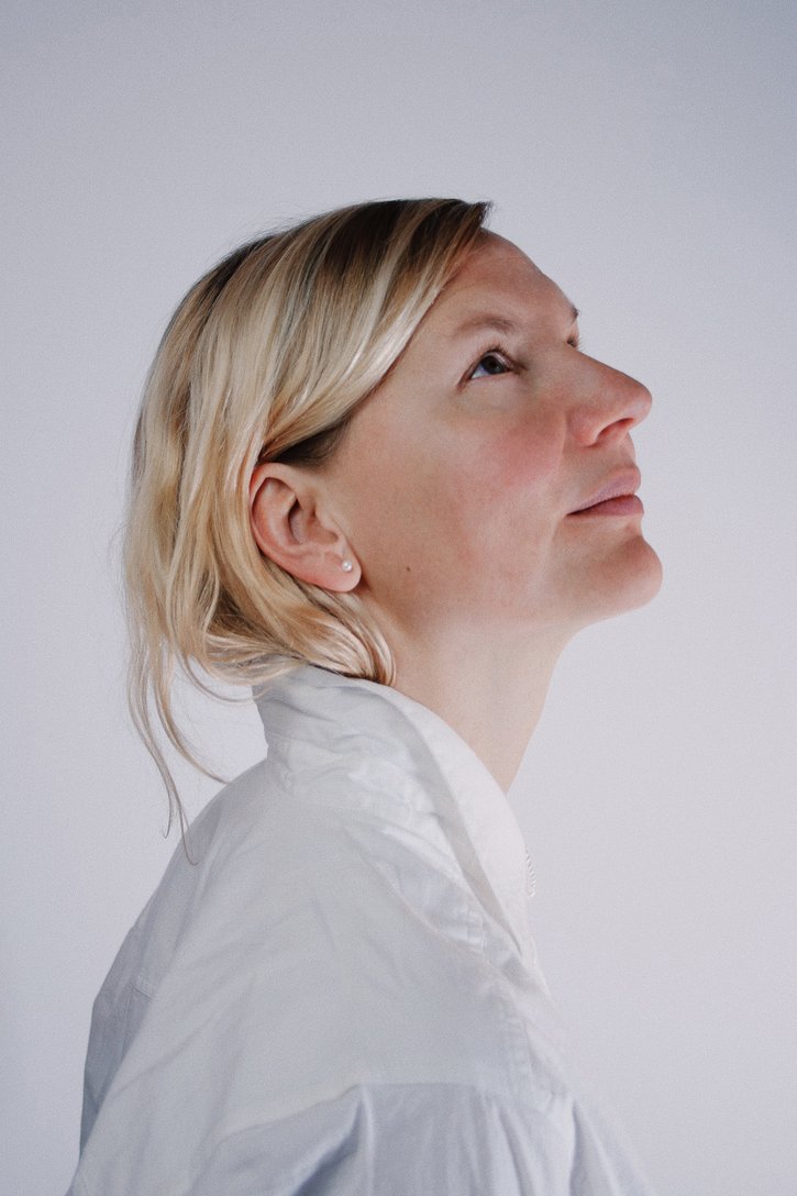 En blond kvinde i en hvid skjorte kigger opad i et lyst rum. Et enkelt ørestykke ses i hendes øre.