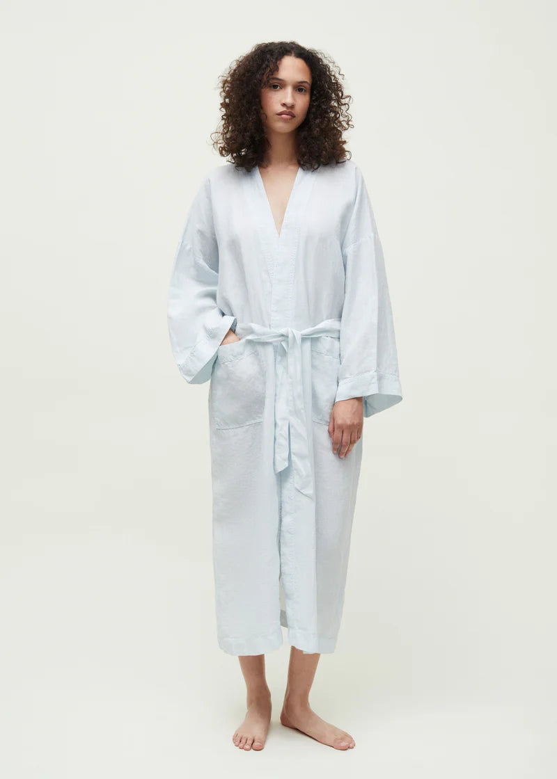 En kvinde står, iført en lyseblå kimono-morgenkåbe med bindebånd. Hun står på en lysegrå baggrund.