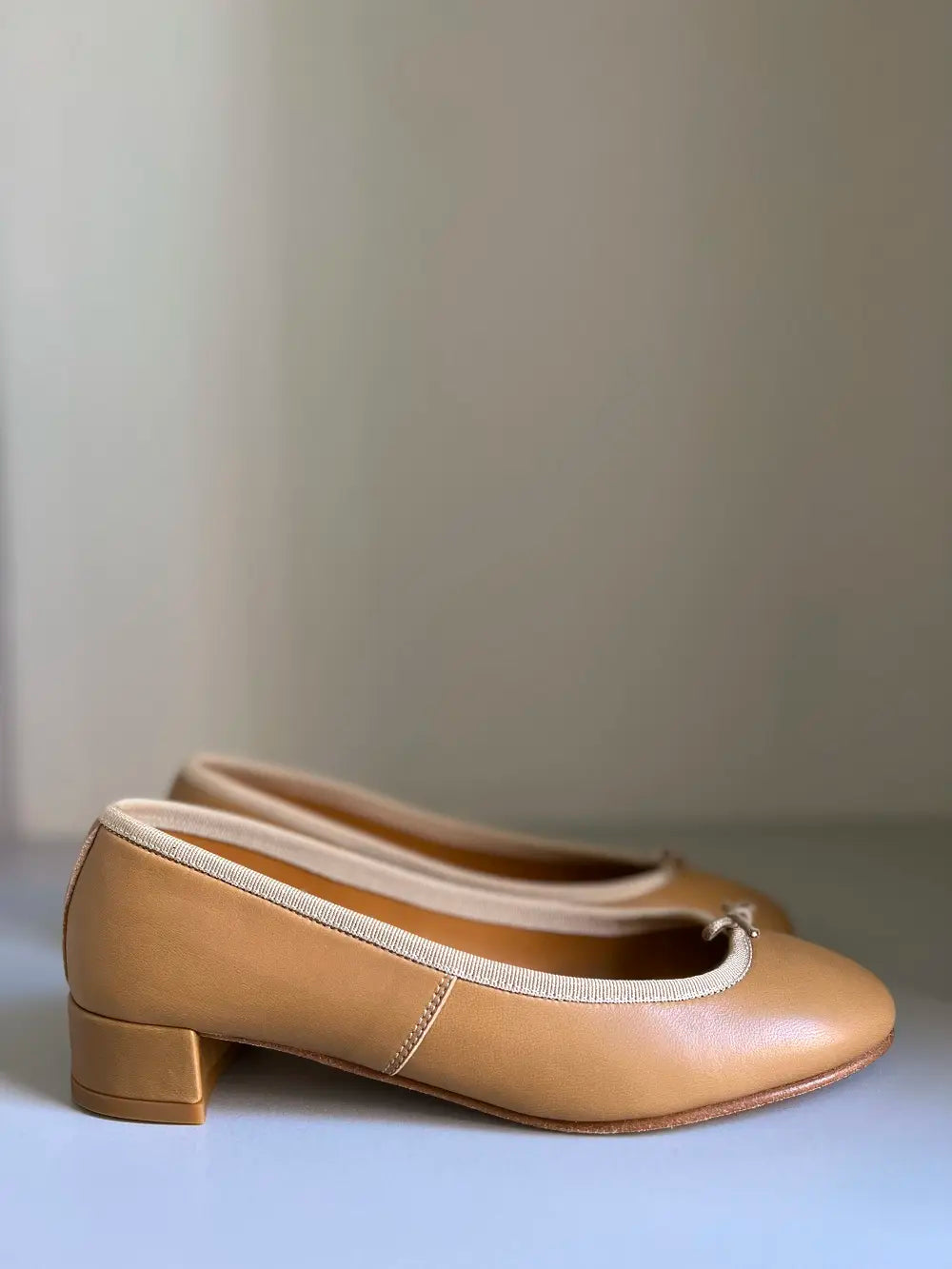 Et par karamelfarvede ballerinasko står side om side på en hvid overflade med en enkel lys beige baggrund.