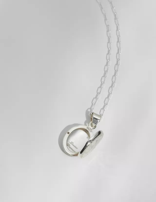 Et sølvfarvet halskædevedhæng, åbent, hænger fra en kæde over en hvid overflade. Den indvendige ring af vedhænget viser inskriptionen "Louise Damas".