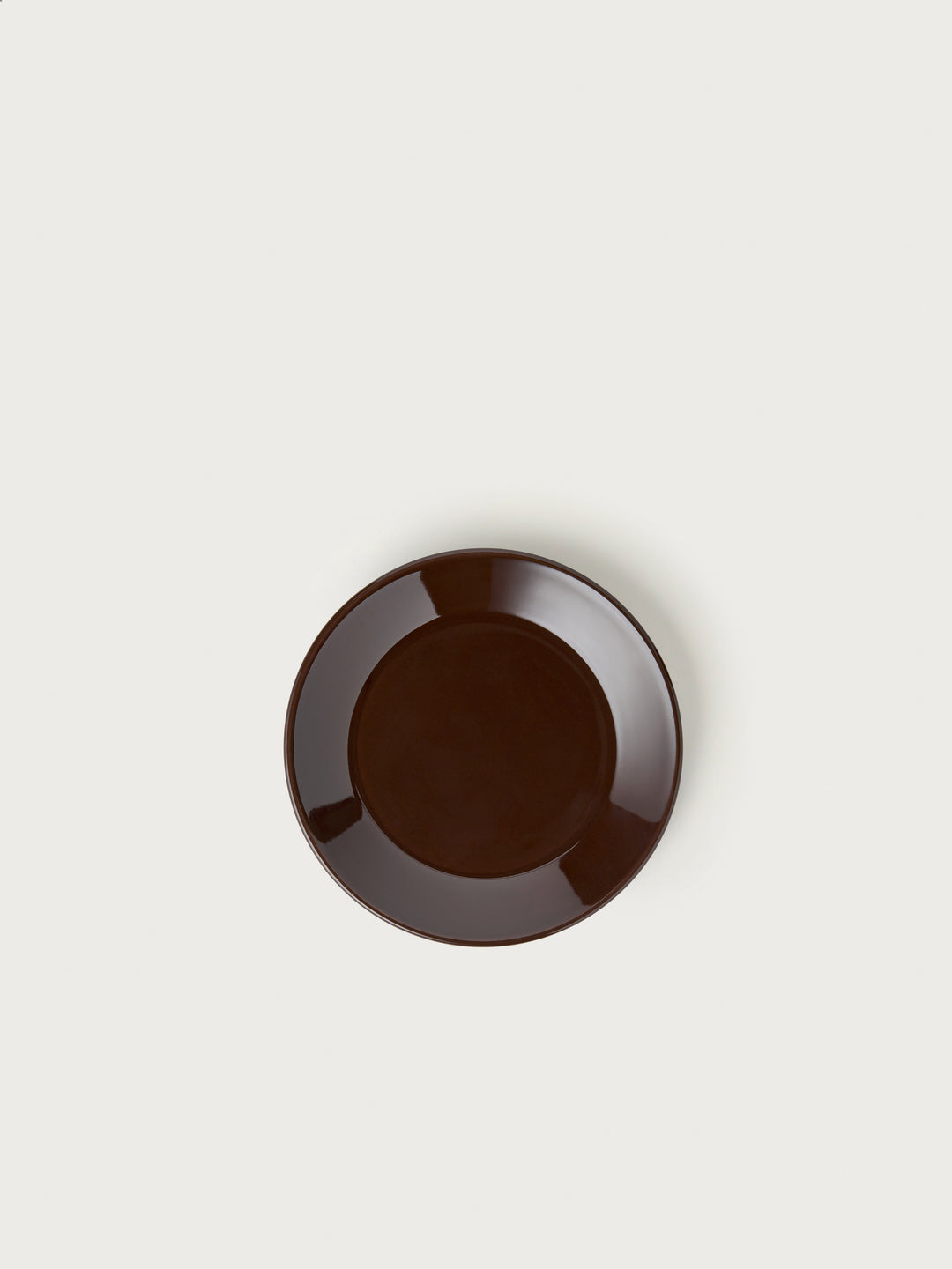 En mørkebrun tallerken hviler centreret på en ensfarvet, cremefarvet baggrund. Tallerkenen reflekterer lyset let.