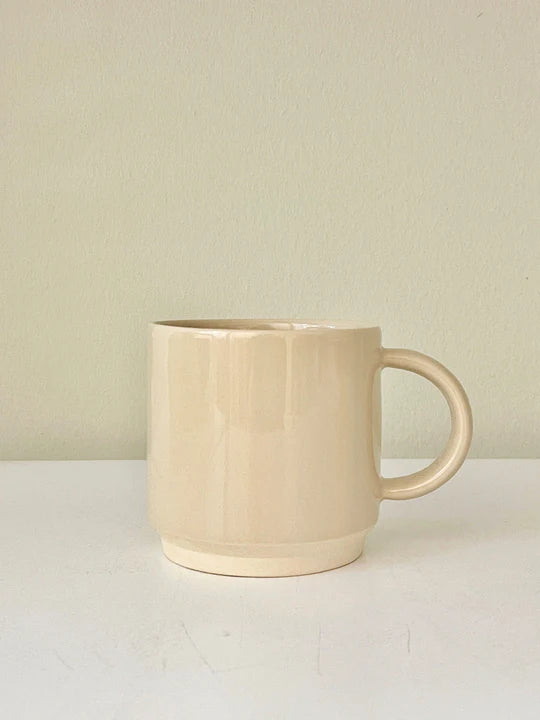 En beige keramisk kop står stille på en hvid overflade foran en ensfarvet beige væg. Koppen er cylindrisk med et afrundet håndtag.