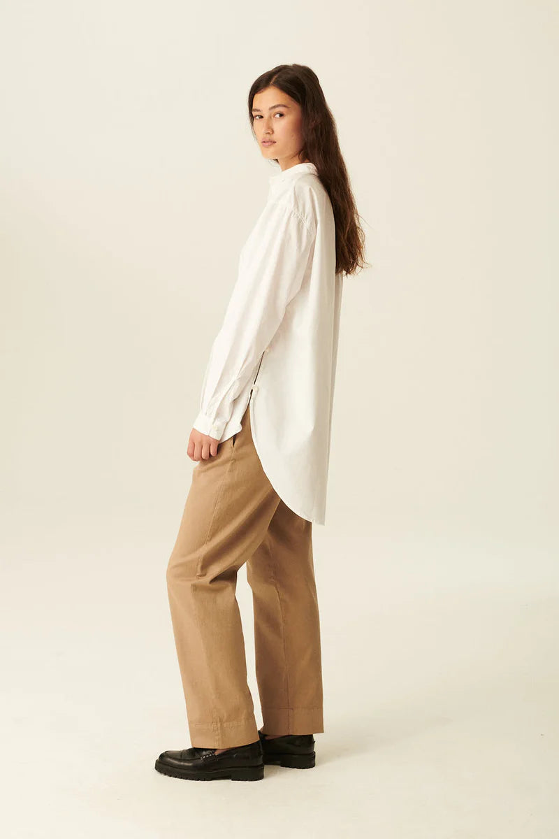 En kvinde i en hvid skjorte og brune bukser står sideværts og kigger over skulderen i et lyst studie.