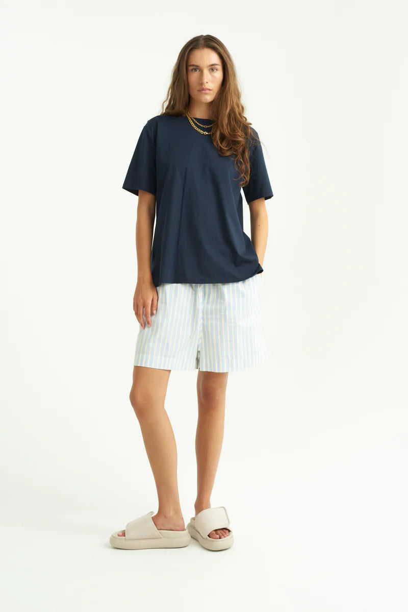 En kvinde står iført en marineblå T-shirt, stribede lyseblå og hvide shorts, og beige sandaler, med hænderne i lommerne, i et hvidt studie.