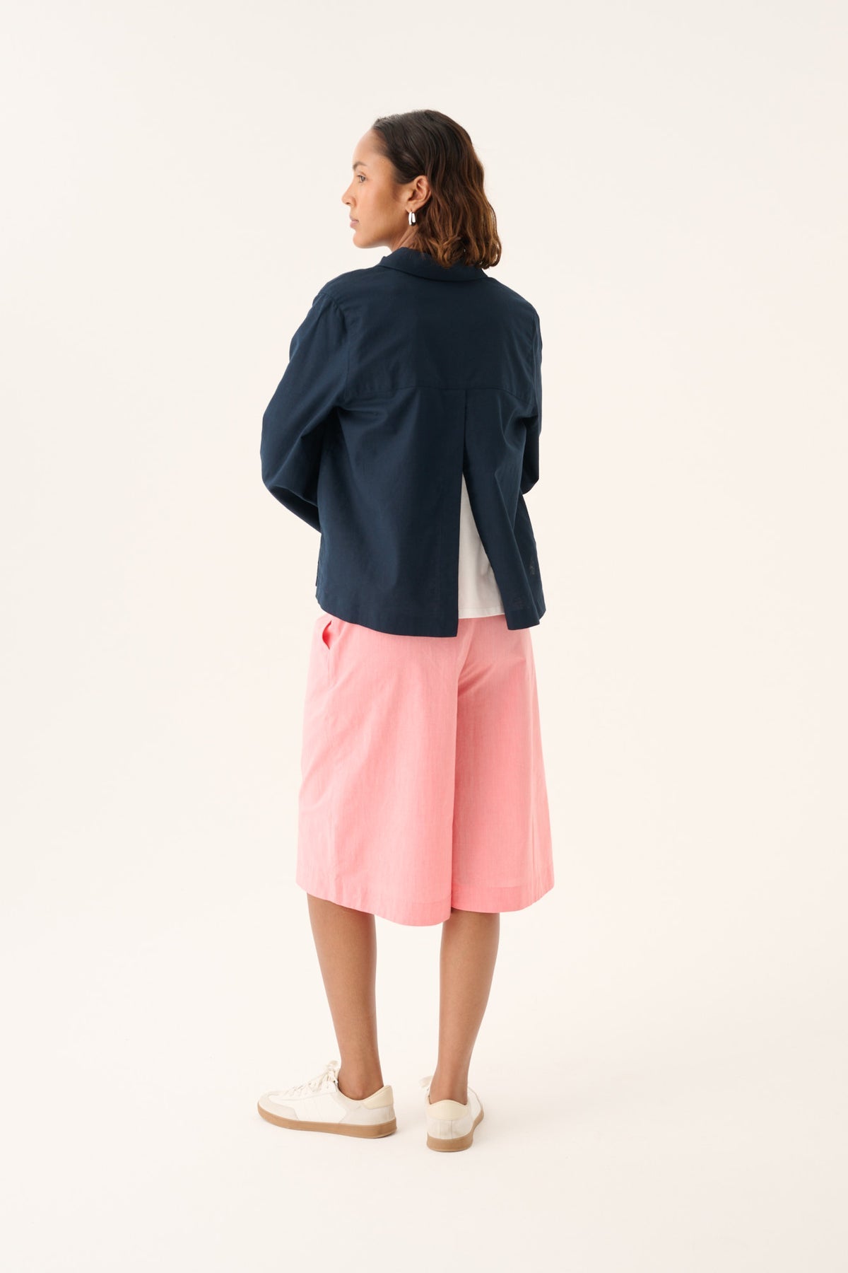 En kvinde står med korslagte arme, iført en mørkeblå jakke og lyserøde shorts. Hun kigger til venstre, med en lys beige baggrund.