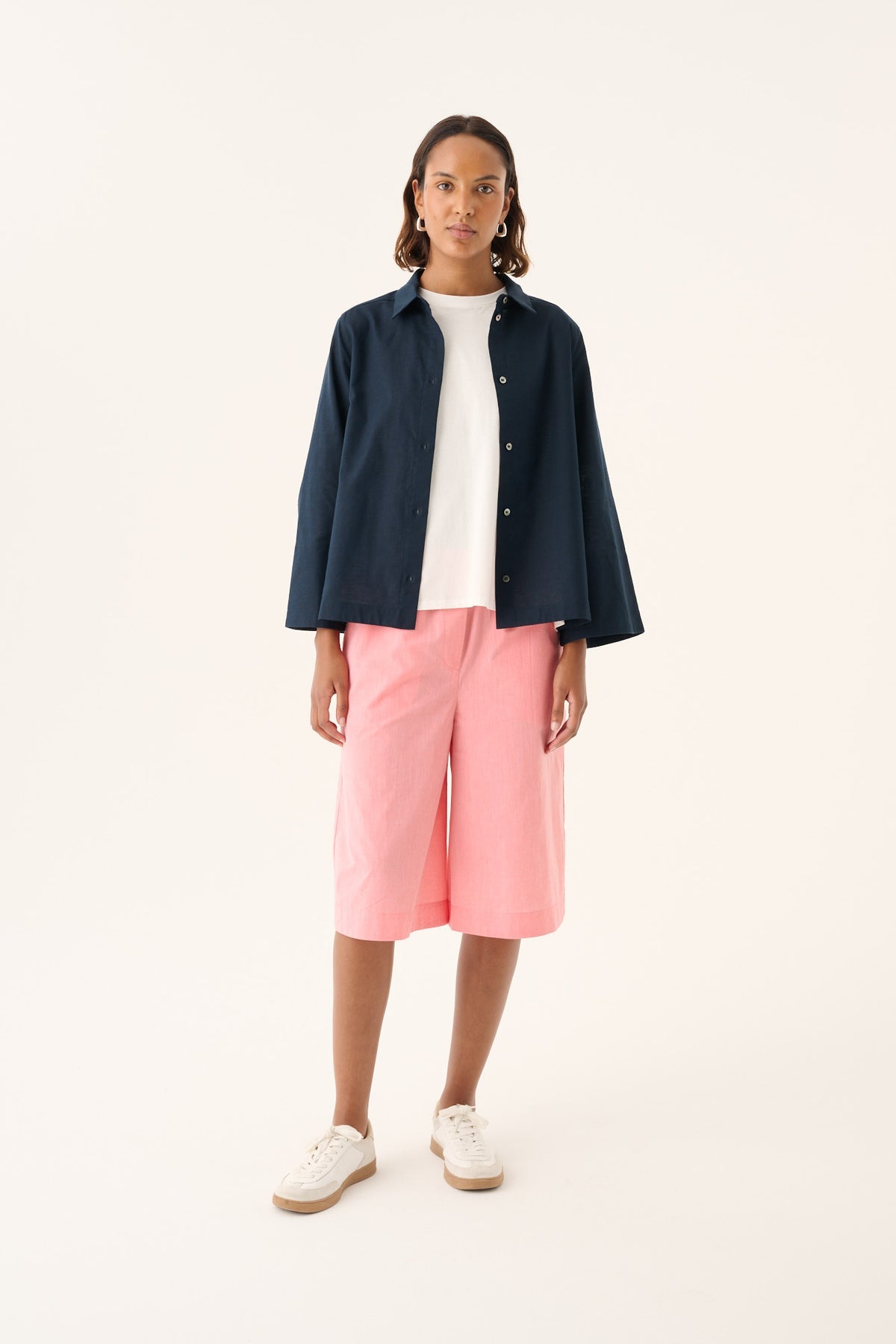 En model står i et studiemiljø iført en marineblå skjorte, en hvid t-shirt og lyserøde shorts, ledsaget af hvide sneakers.